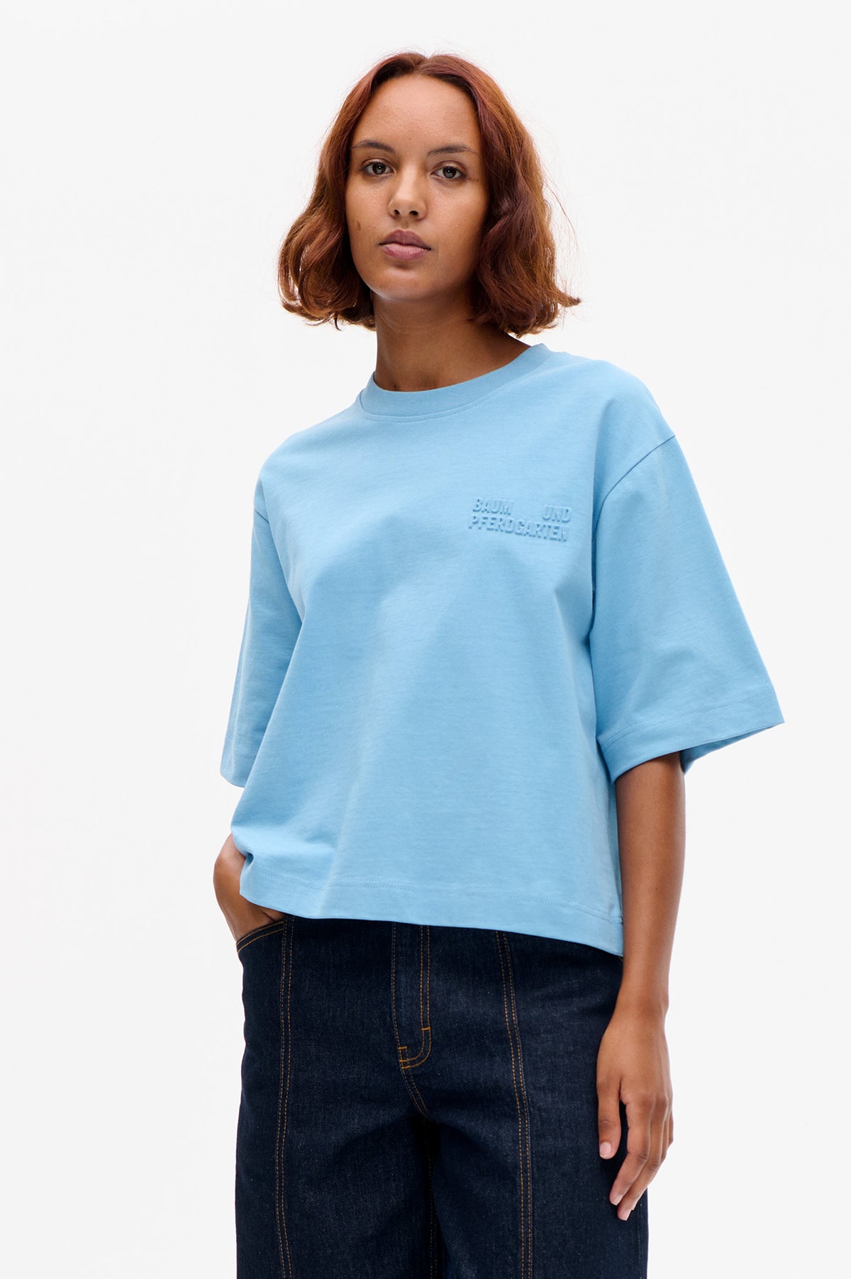 Vue de face d'un mannequin portant un t-shirt bleu clair à manches courtes, avec un logo ton sur ton en relief sur la poitrine et un jean foncé.
