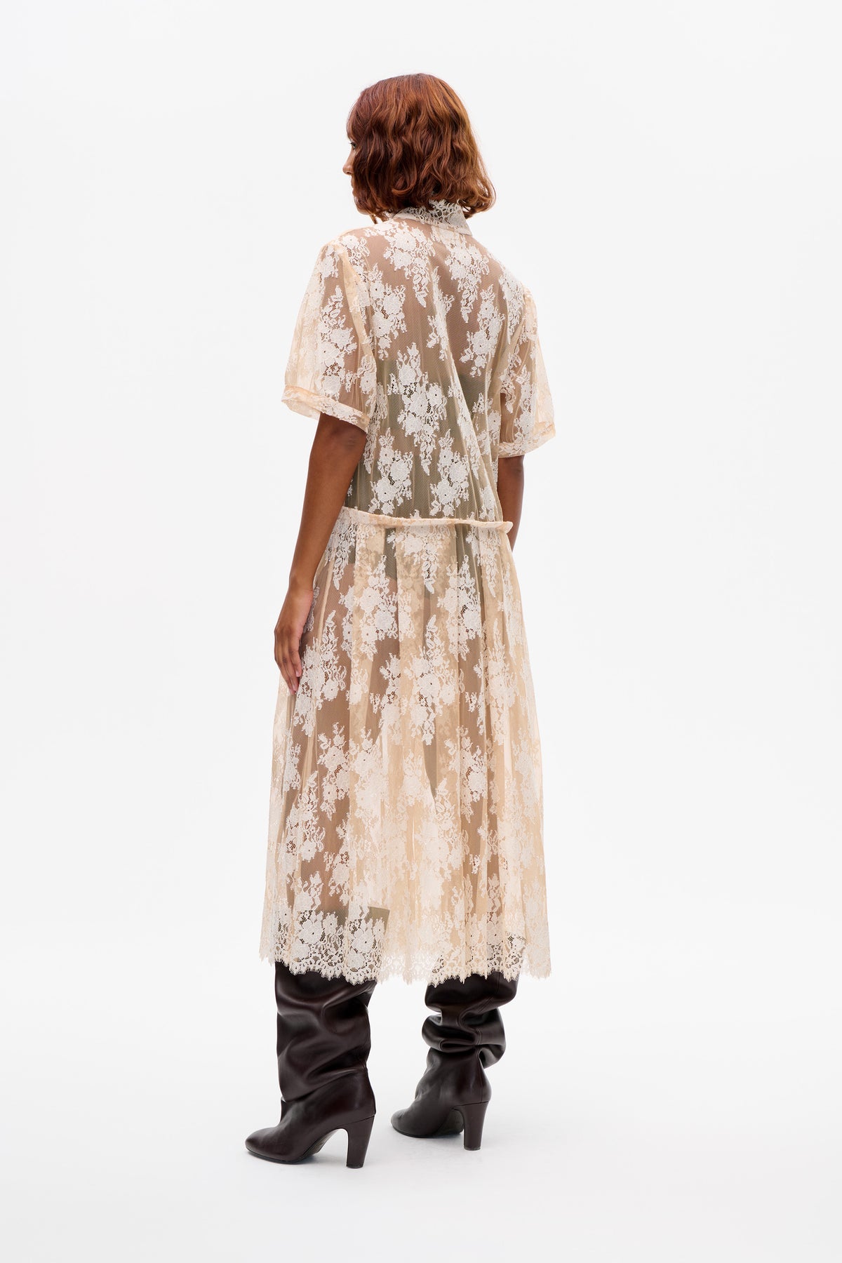 Vue de dos d'un mannequin portant une robe en dentelle florale ivoire transparente à manches courtes et col montant, portée sur un pantalon foncé.