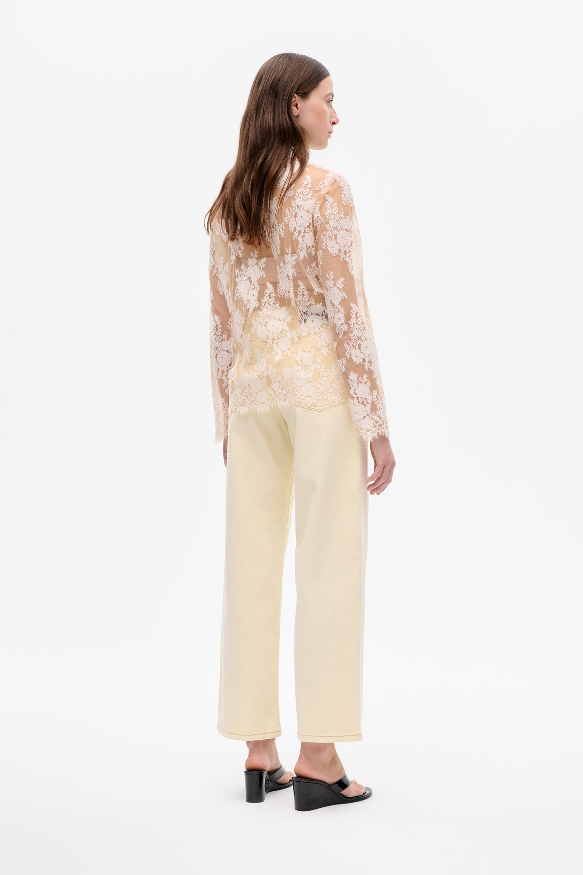 Seitliche Rückansicht eines Models, das eine transparente pfirsichfarbene Bluse mit weißer Blumen-Spitze und eine cremefarbene, weit geschnittene, knöchellange Hose trägt.