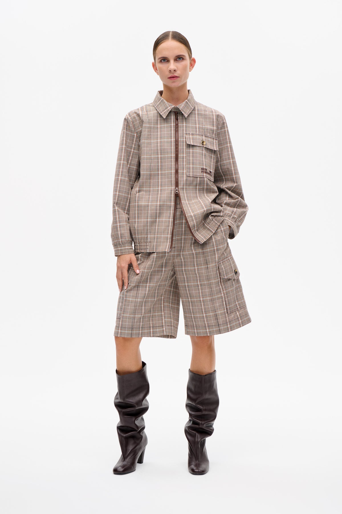 Model wearing a beige and brown plaid zip-front jacket with a flap chest pocket and matching knee-length shorts, styled with dark knee-high boots.