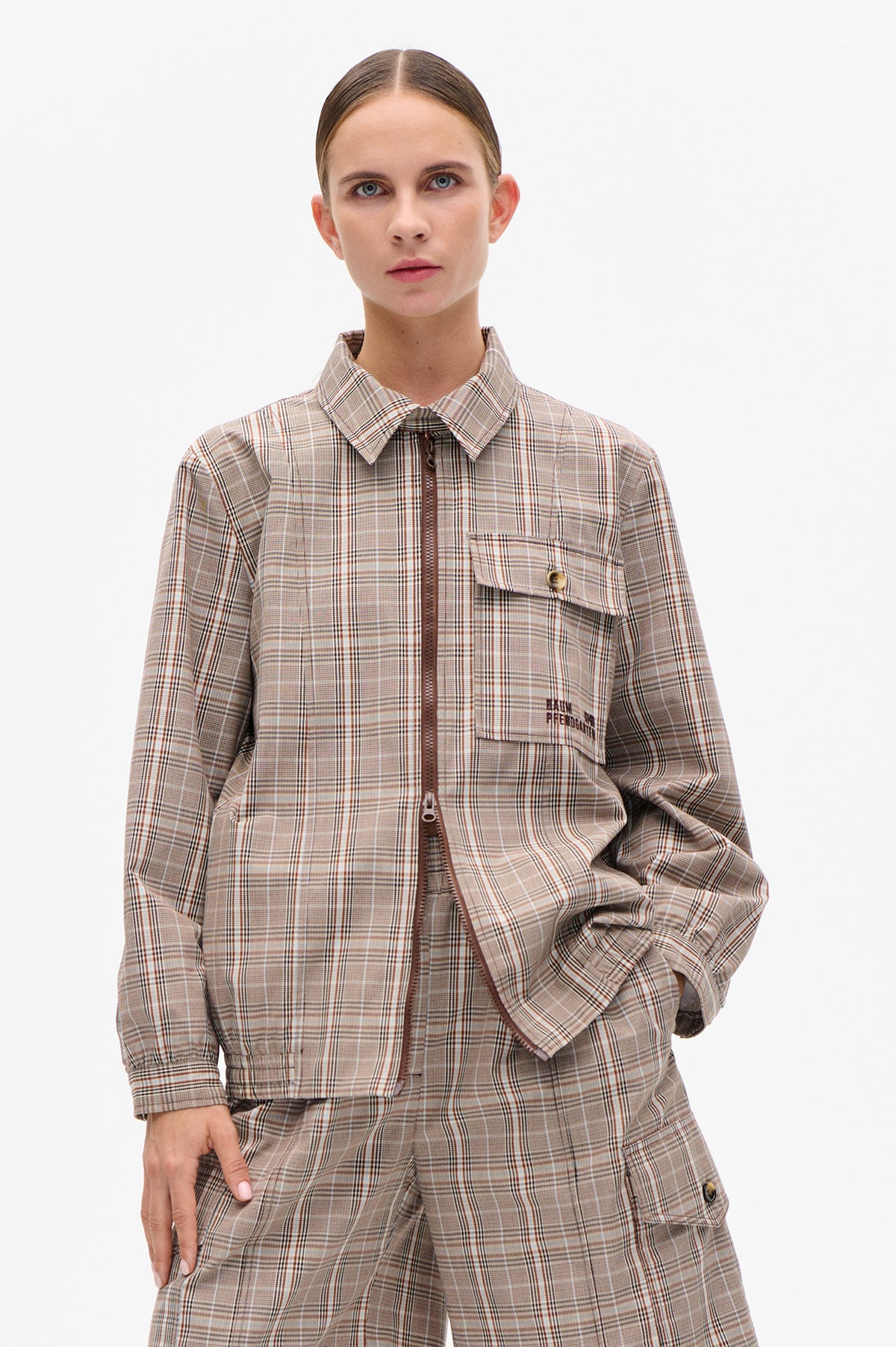 Front view of a model wearing a taupe plaid zip-front jacket with a pointed collar and flap chest pocket, paired with matching trousers.