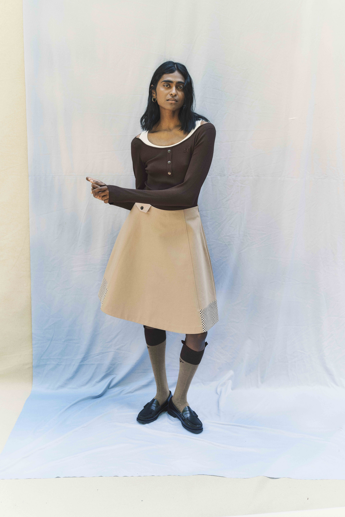 Model wearing a dark brown ribbed button-front cardigan with a cream neckline and beige skirt with checked panels against a pale blue backdrop.