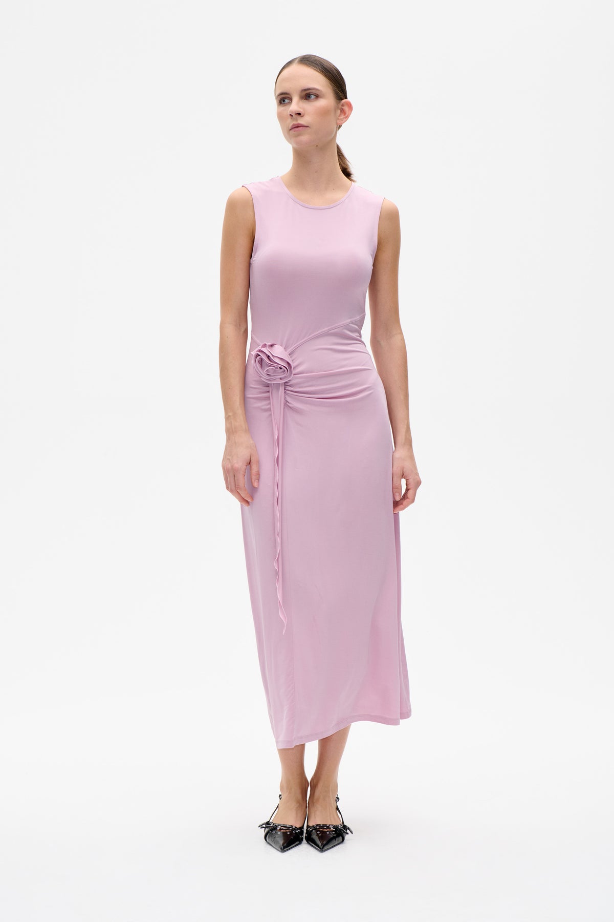 Front view of a model wearing a sleeveless light pink midi dress with a round neckline and gathered side draping with a fabric rosette.