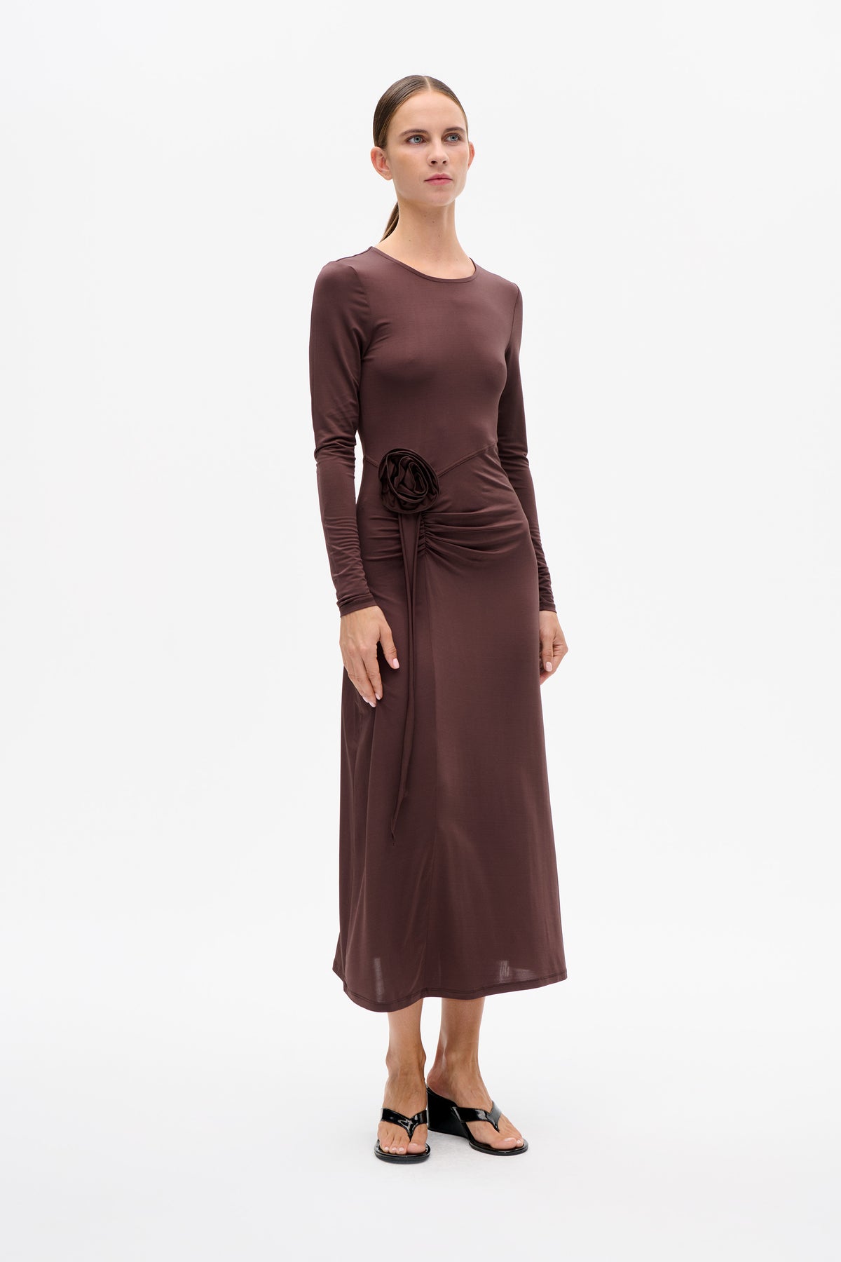Front view of a model wearing a long-sleeved plum-brown dress with a wide neckline, gathered waist and fabric rosette detail.
