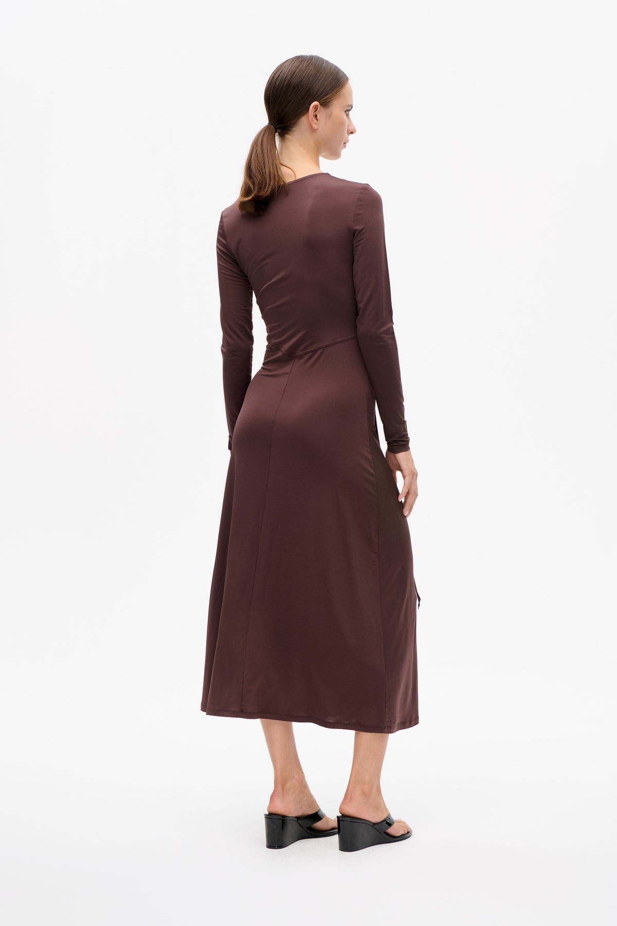 Achteraanzicht van een model in een effen diepbruine midi-jurk met lange mouwen, een getailleerde pasvorm en een schuine naad.