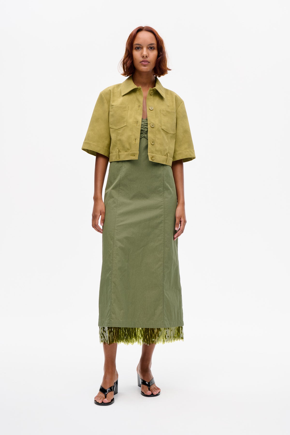 Front view of a model in a yellow-olive cropped jacket with short sleeves and patch pockets over a sage green dress with a sequinned hem.