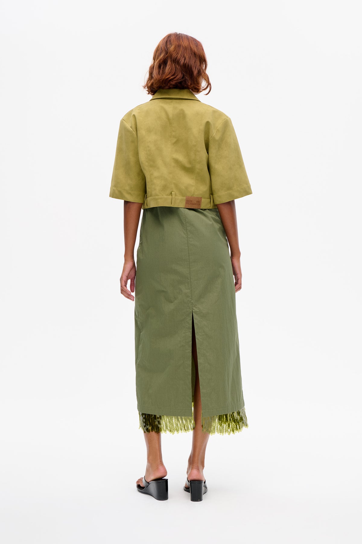 Back view of a model wearing a cropped olive-green short-sleeved jacket with a pointed collar and waist tabs over a green dress with a sequined hem.