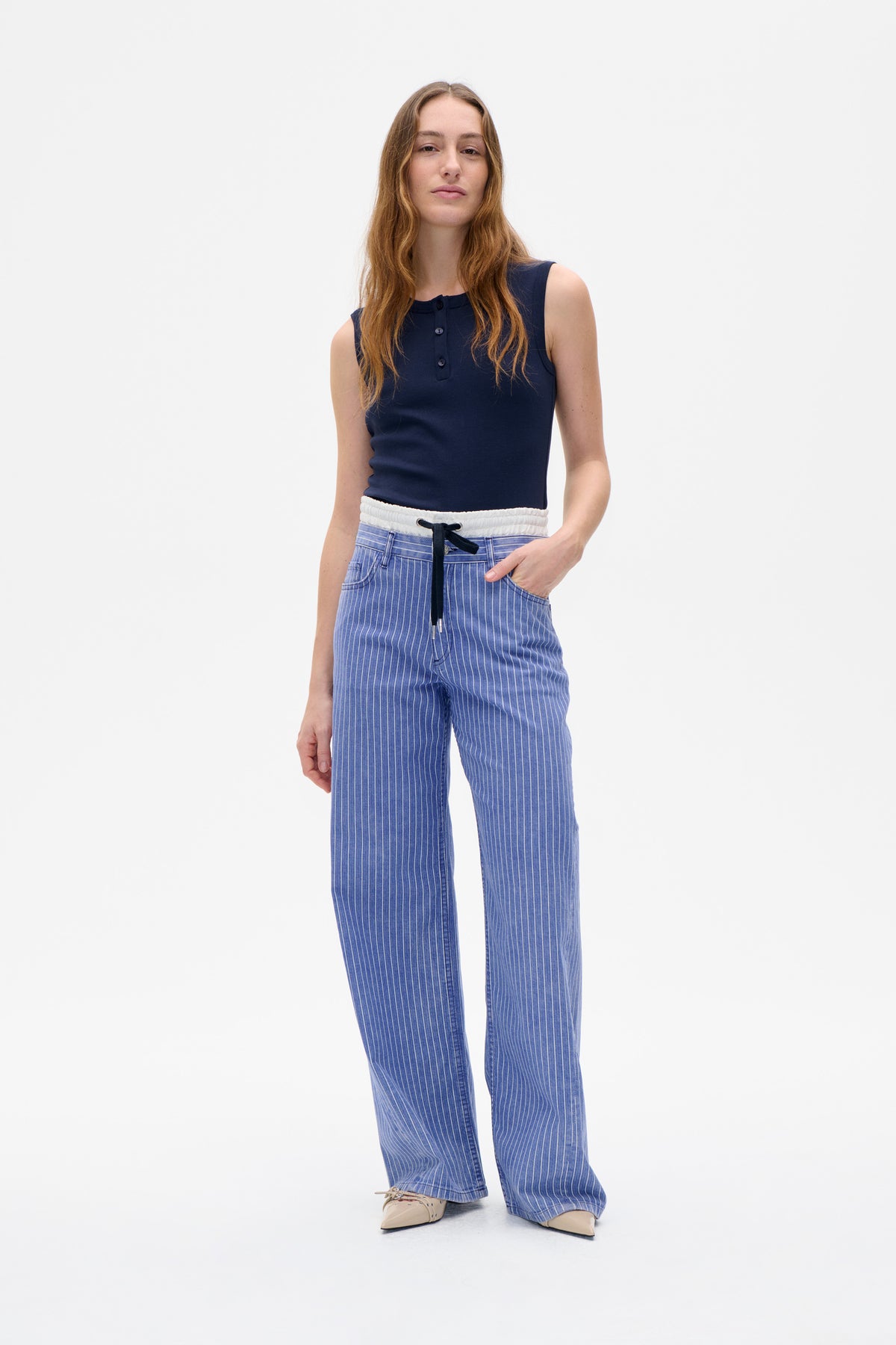 Model trägt ein marineblaues ärmelloses Top mit Knopfleiste und eine blaue, weit geschnittene Hose mit feinen weißen Nadelstreifen und einem Kordelzug in der Taille.