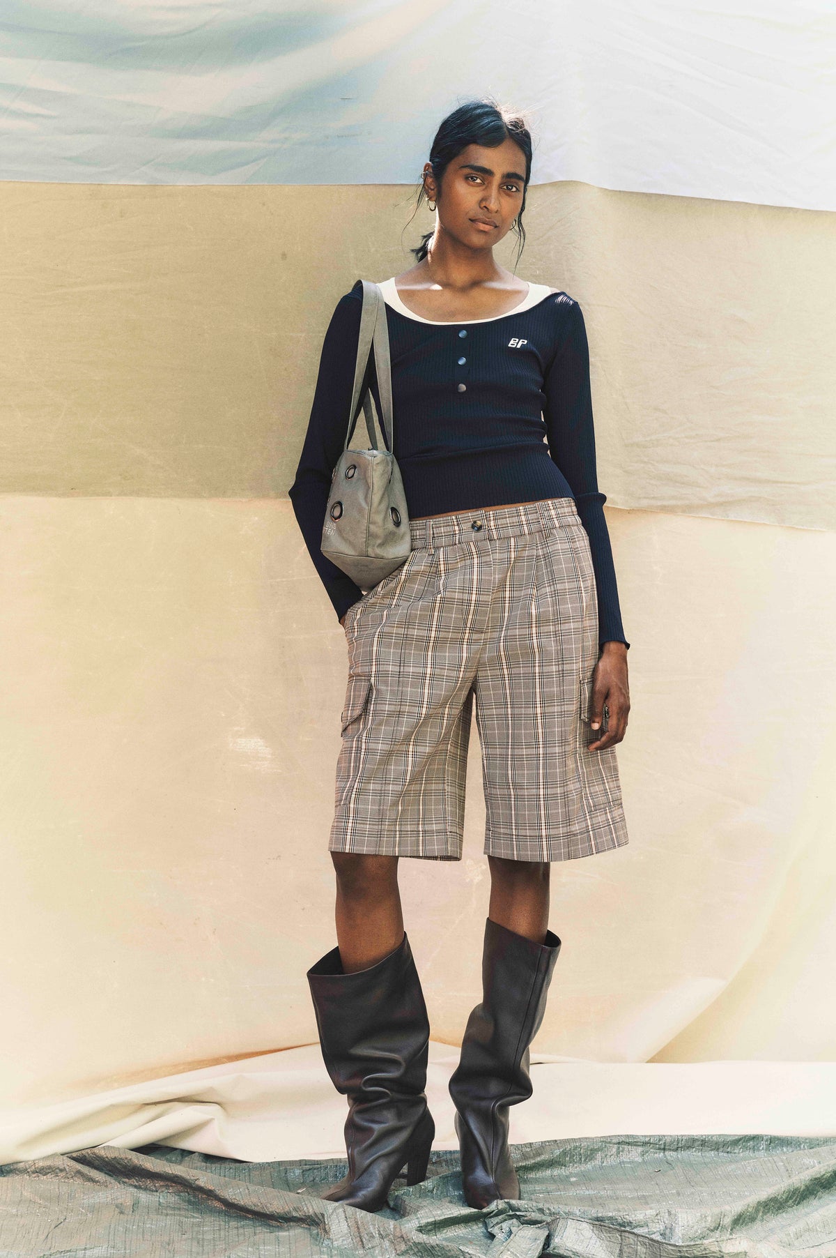 Campaign model wearing a navy ribbed long-sleeve top with white neckline, beige plaid shorts, a gray shoulder bag and black boots
