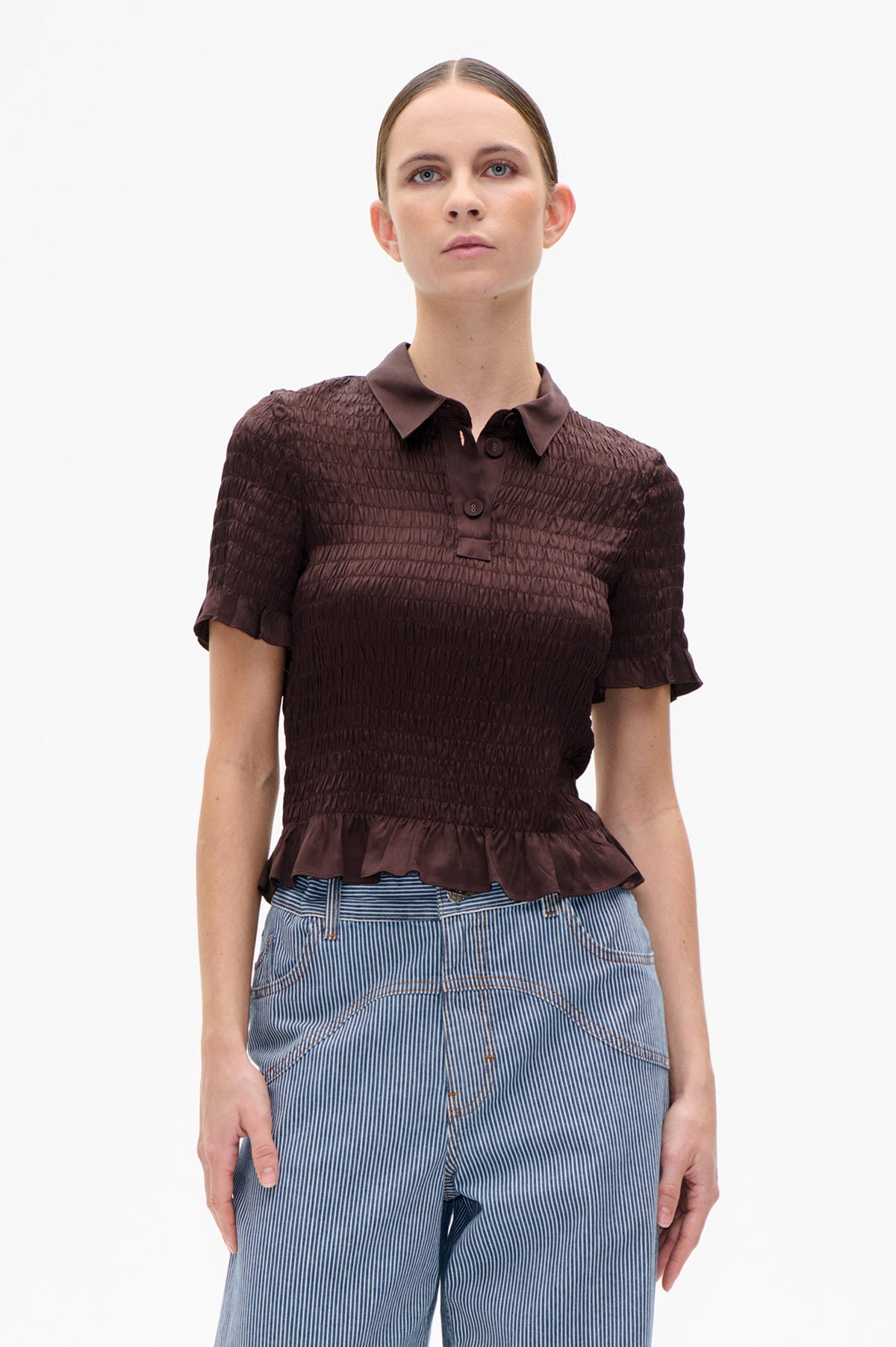 Front view of a dark brown short-sleeved collared blouse with gathered horizontal texture and a flared hem, paired with blue-and-white striped jeans.