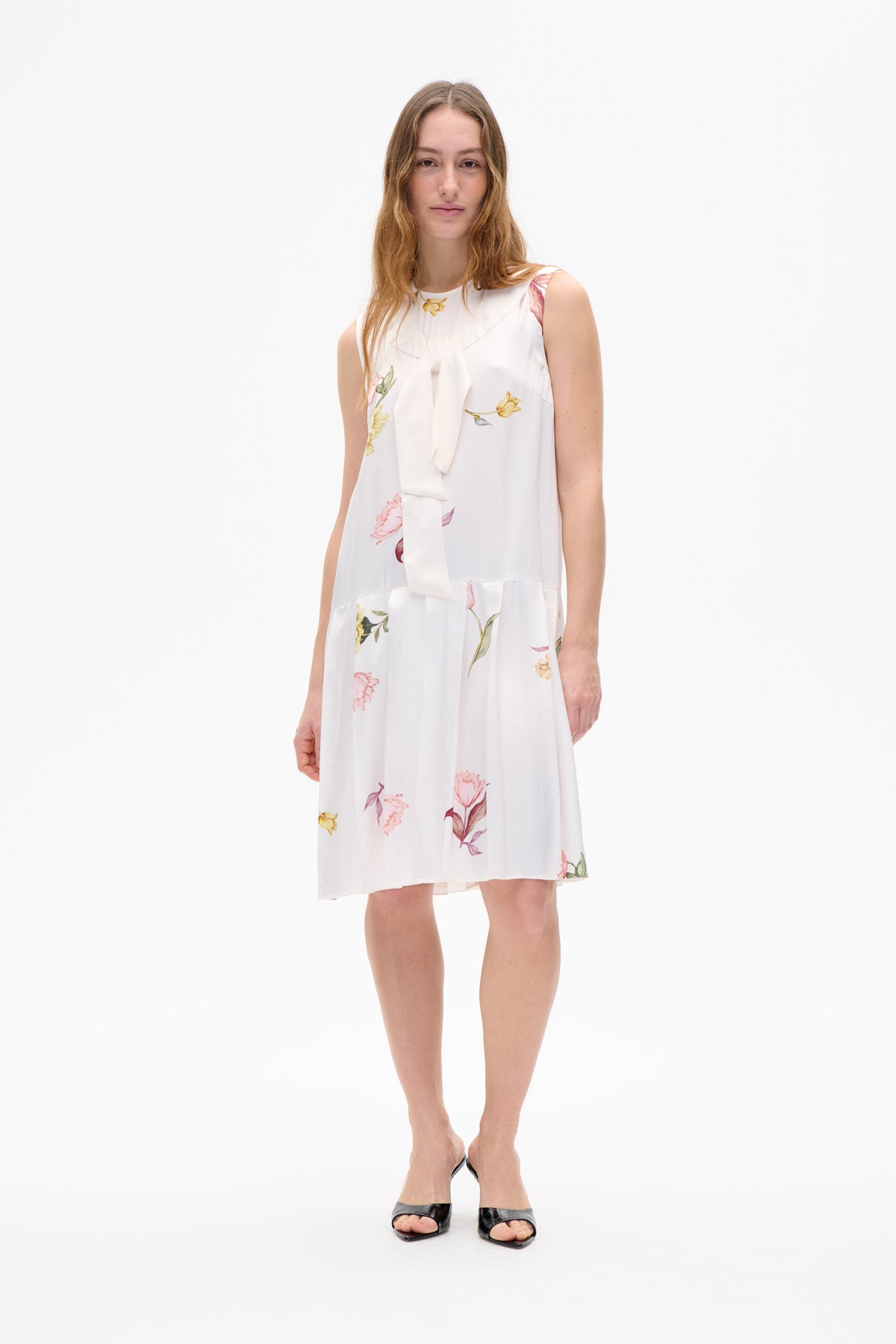 Model wearing a sleeveless white dress with a long neck tie and scattered pink and yellow floral print, shown from the front.