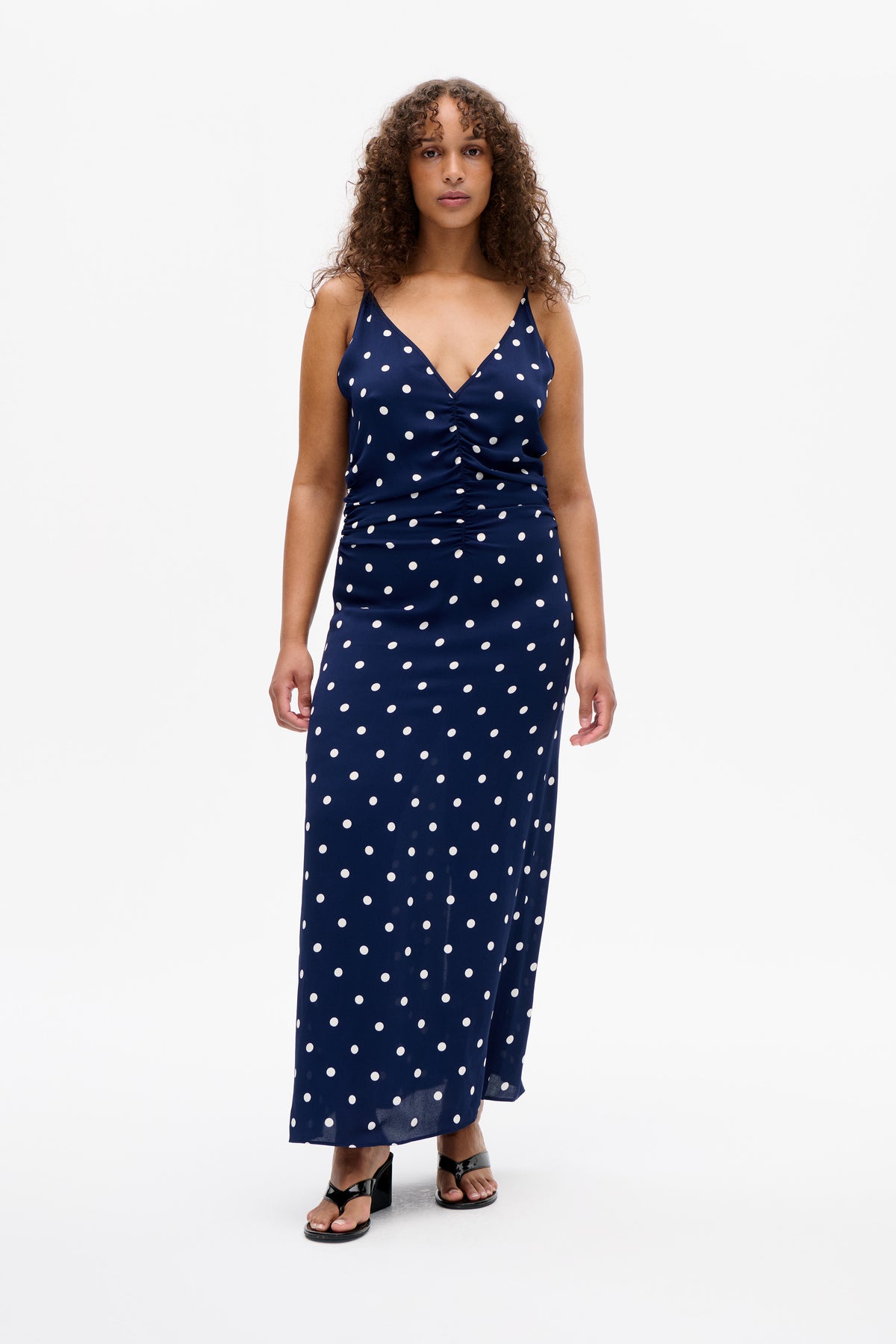 Front view of a model wearing a navy spaghetti-strap midi dress with white polka dots, a V-neckline and gathered front.