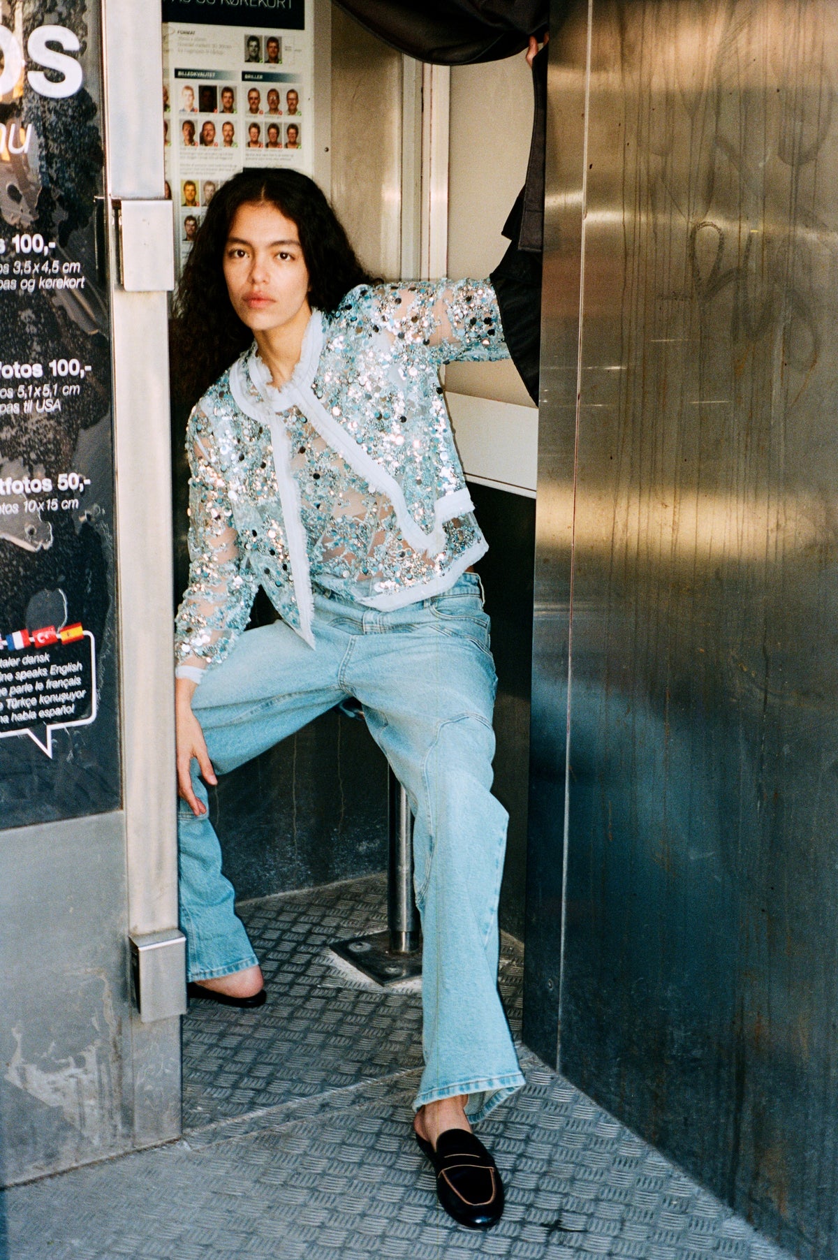 Campagneportret van een zittend model dat een doorschijnende lichtblauwe blouse met subtiele versieringen, een lichte gerimpelde kraag en lichtgewassen jeans draagt.