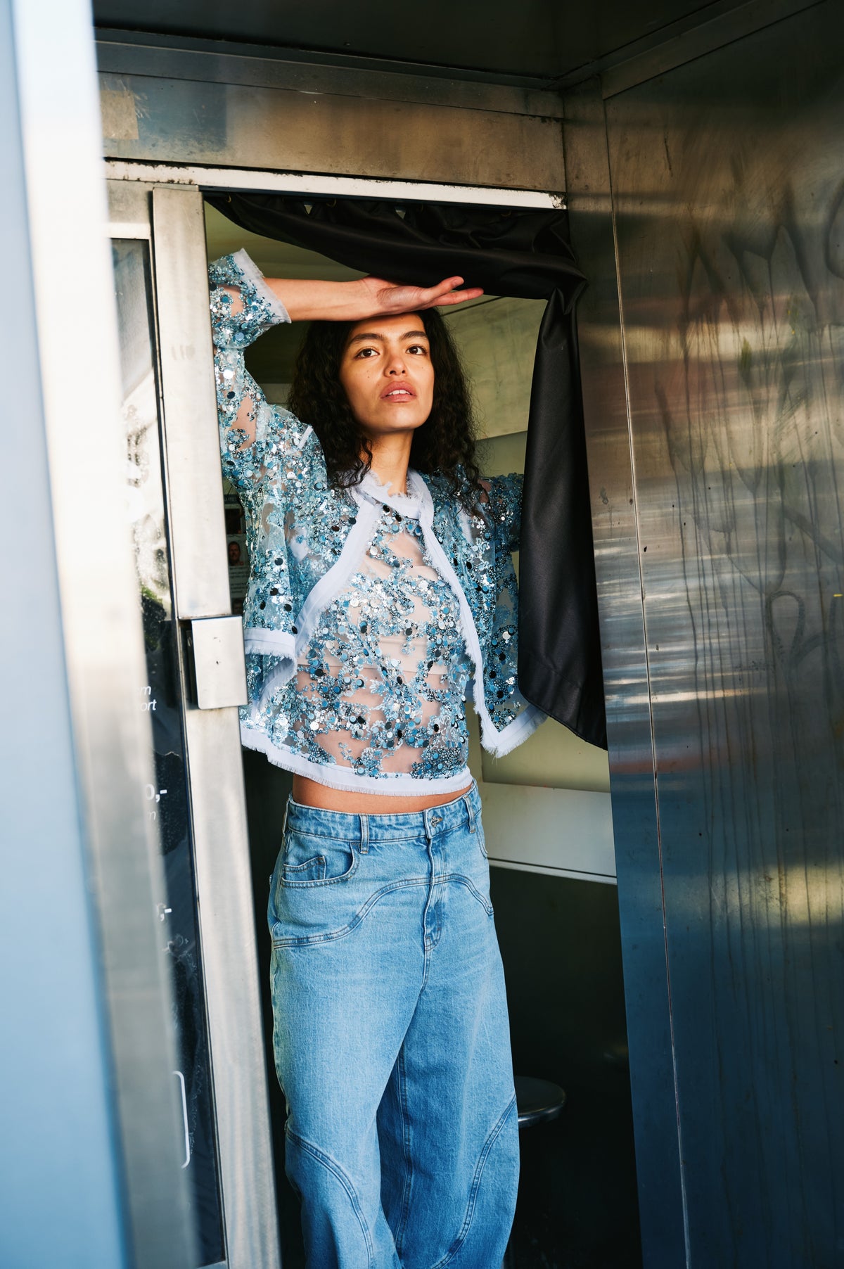 Campaign portrait of a model lifting a black curtain, wearing a sheer pale blue top embellished with blue and silver sequins and light-wash jeans.