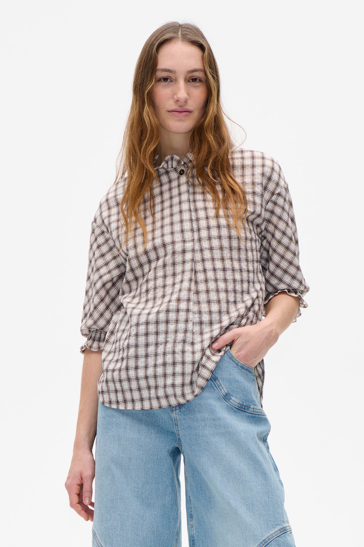 Model draagt een crèmekleurige, geruite blouse met knopen aan de voorkant, ruches bij de hals en gerimpelde mouwen, gecombineerd met lichtblauwe jeans.