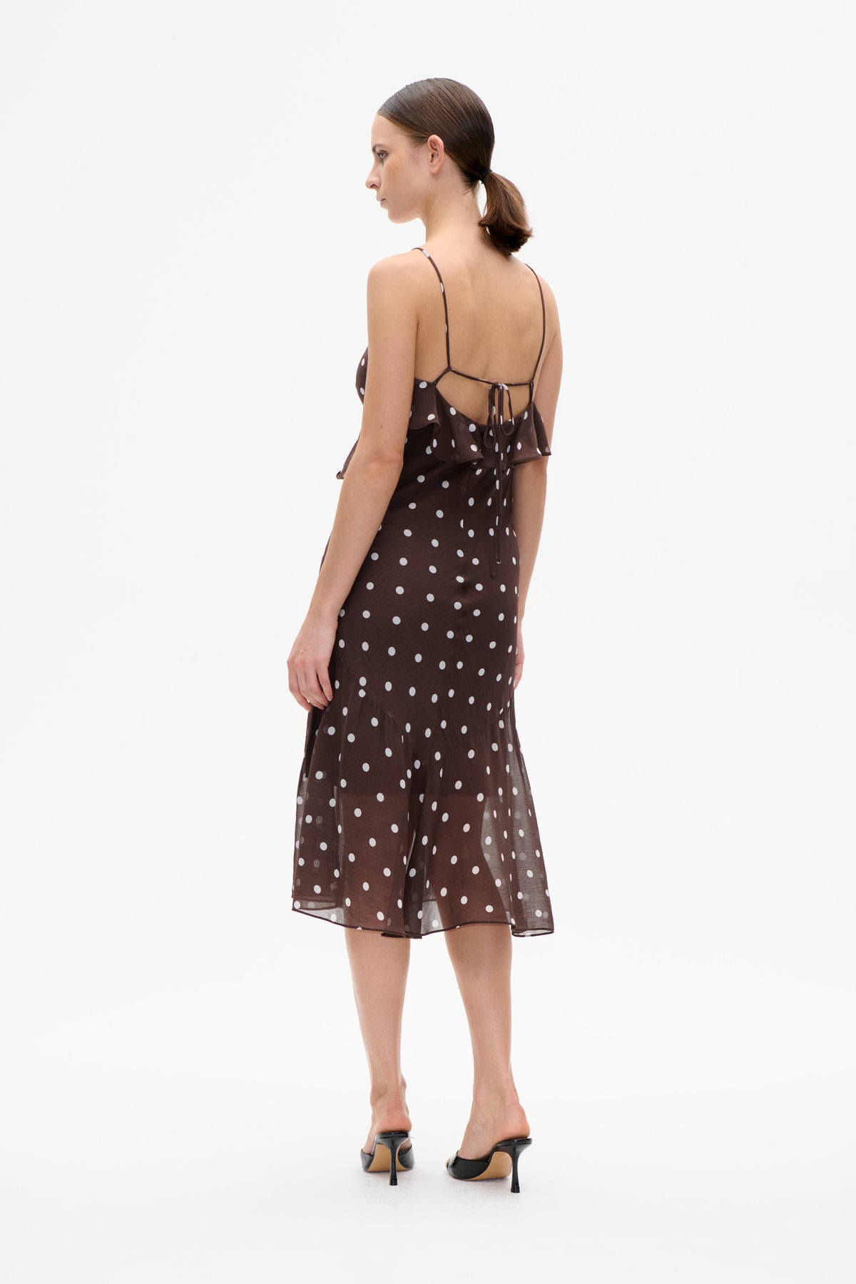 Model wearing a dark brown midi dress with white polka dots, ruffled hem and thin tie-back straps, shown from behind.