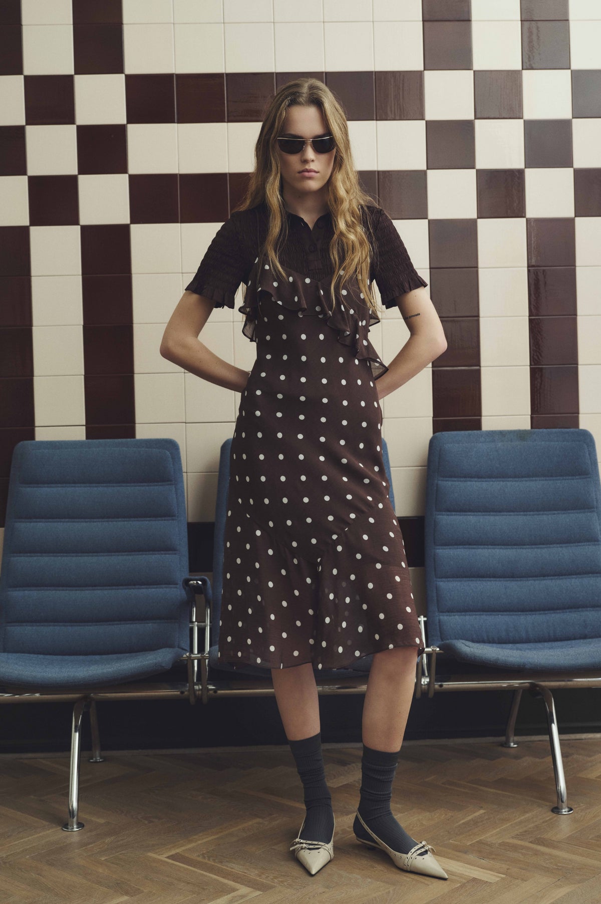Model in a dark brown dress with white polka dots, layered ruffles and short sleeves, posed between blue chairs against a checkerboard tile wall.