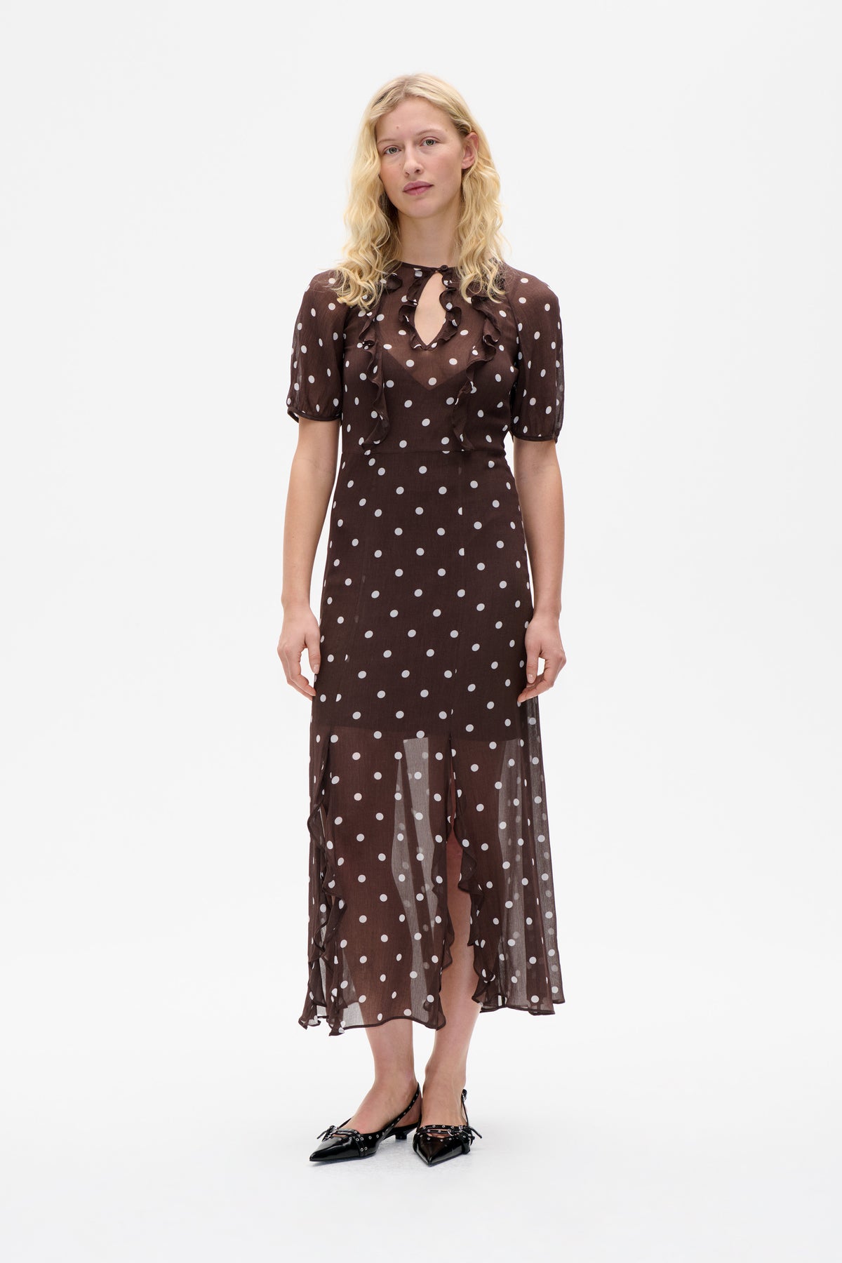 Front view of a model wearing a sheer dark brown midi dress with white polka dots, ruffled neckline and short puff sleeves.