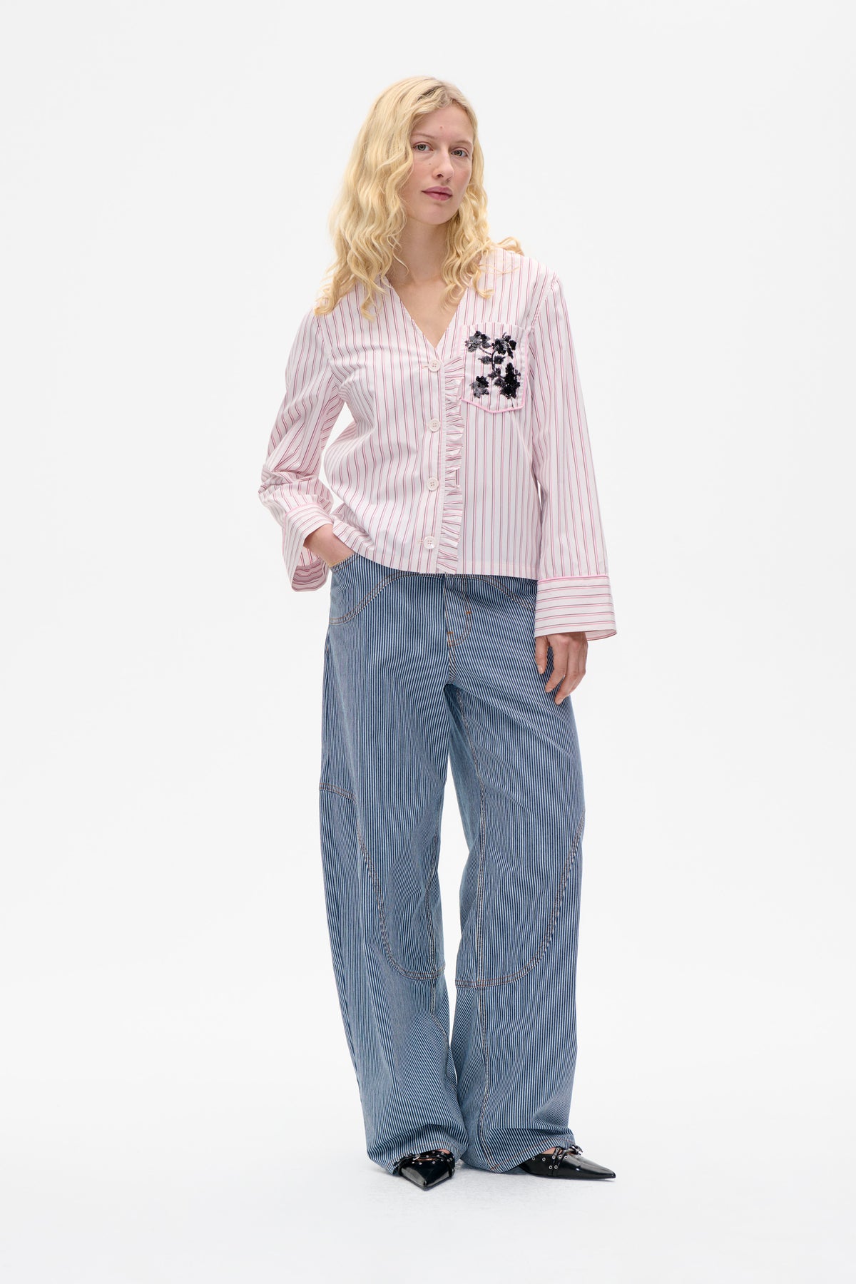 Model wearing a white blouse with pink stripes, ruffled button placket and embellished chest pocket, paired with blue pinstripe trousers.