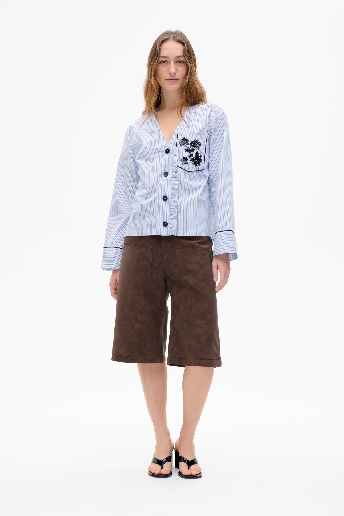Front view of a model wearing a pale blue pinstriped button-front blouse with a ruffled placket and embellished chest pocket, paired with brown knee-length shorts.