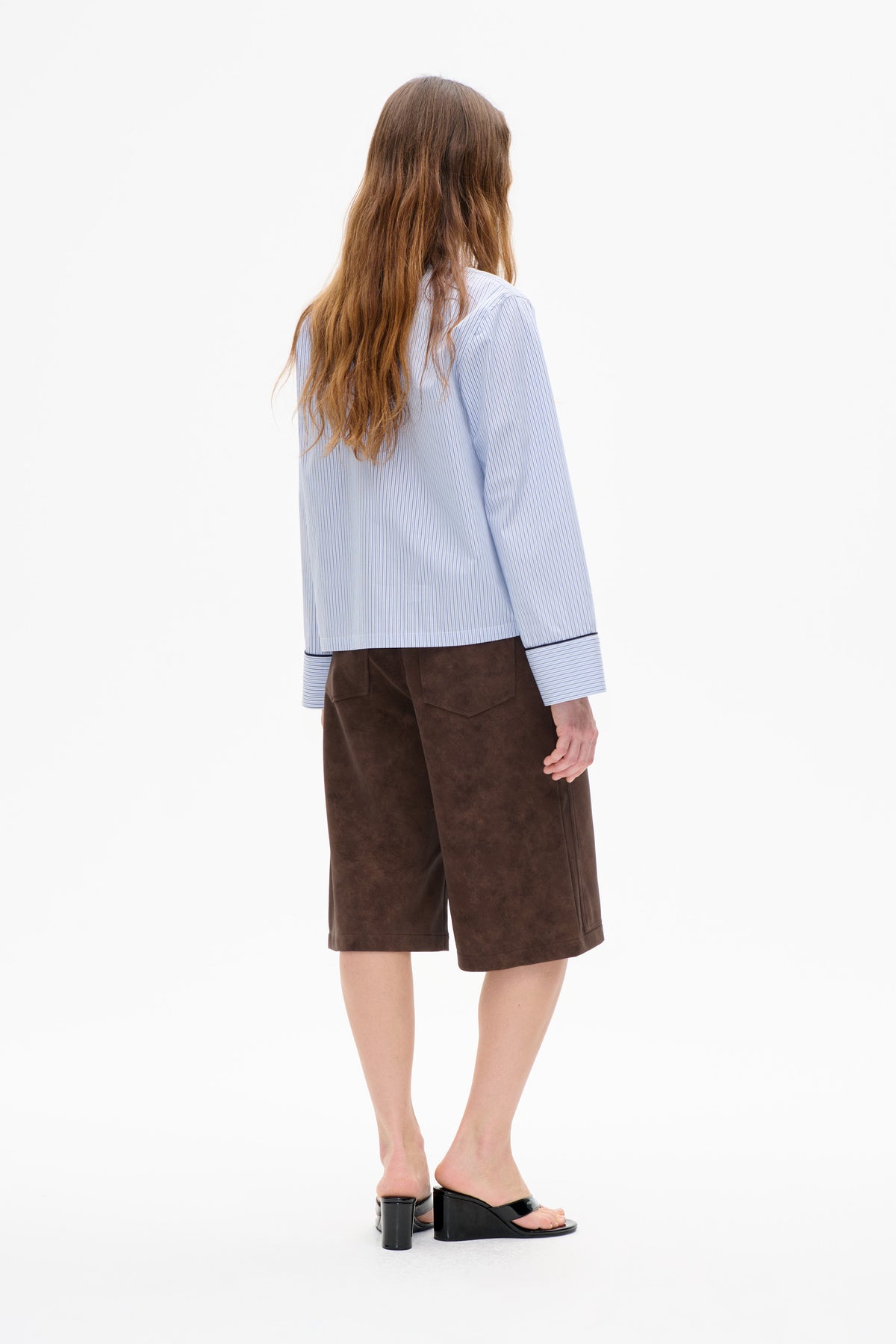 Rear view of a model wearing a pale blue fine-striped long-sleeve shirt with dark cuff trim and brown knee-length shorts.