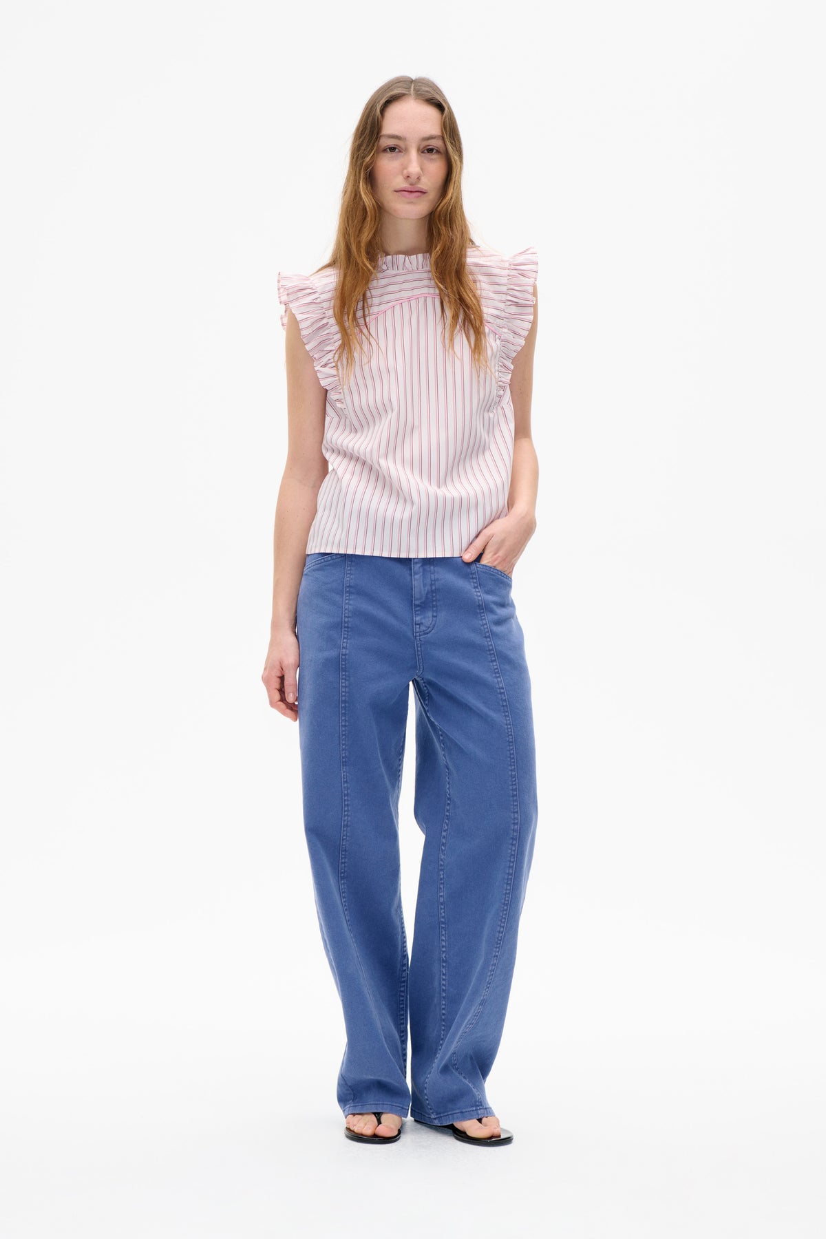 Vooraanzicht van een model dat een witte top met roze verticale strepen draagt, met een hoog geplooide halslijn en ruches op de schouders, gecombineerd met een blauwe broek met wijd uitlopende pijpen.