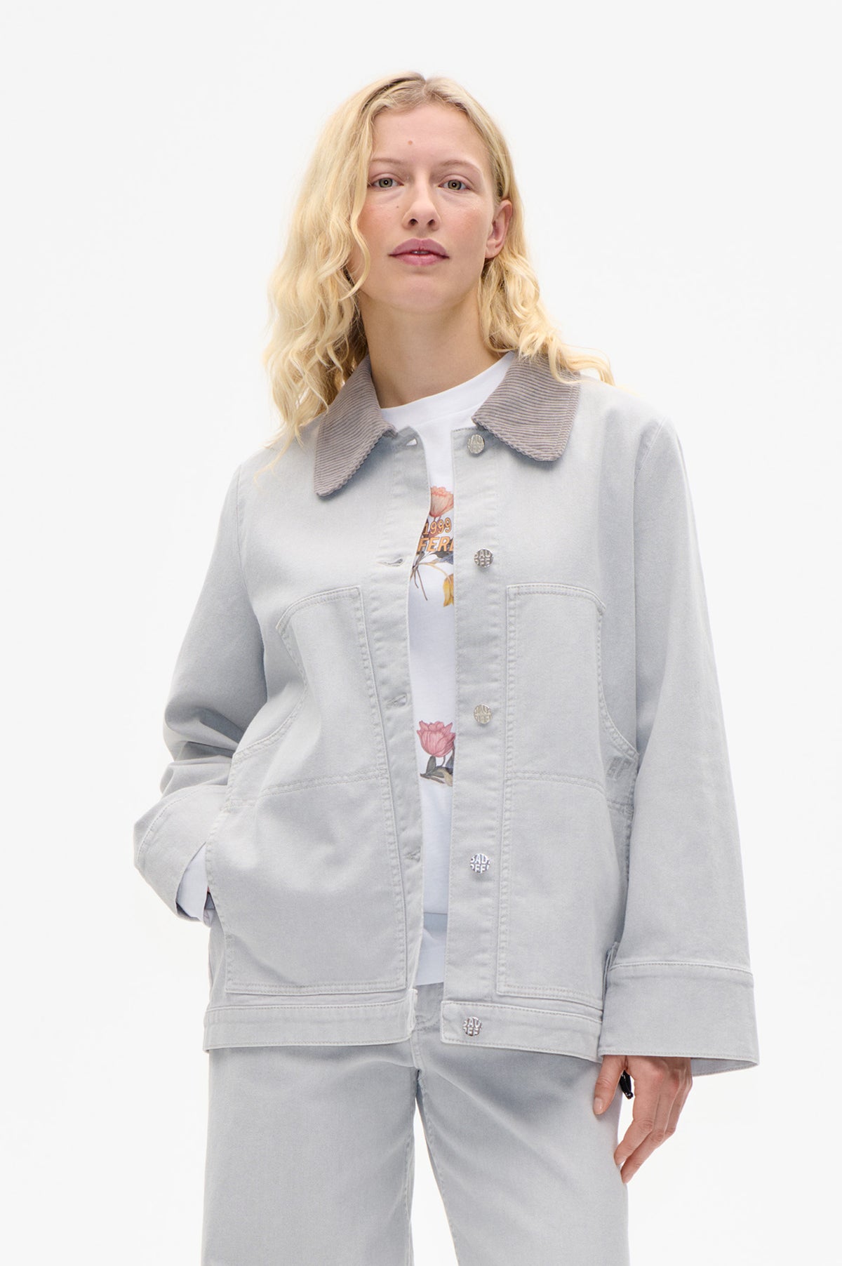 Front view of a model wearing a light grey denim jacket with a striped collar, metal buttons and large patch pockets over a white graphic T-shirt.