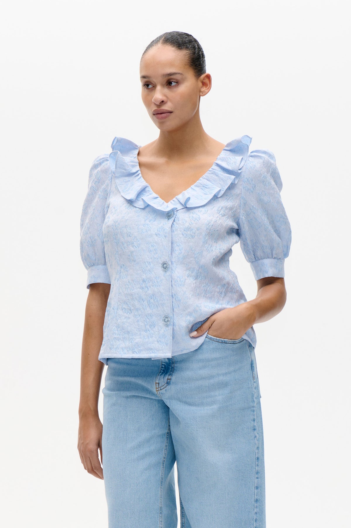 Model wearing a pale blue textured button-front blouse with a ruffled neckline and puff sleeves, paired with light-wash jeans.