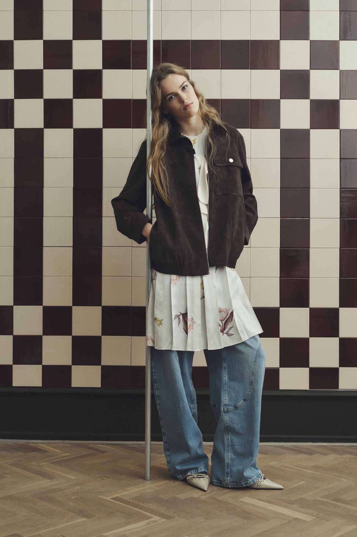 Mannequin portant une veste zippée marron foncé par-dessus une tunique blanche à fleurs et un jean large bleu, posant devant un mur en damier.