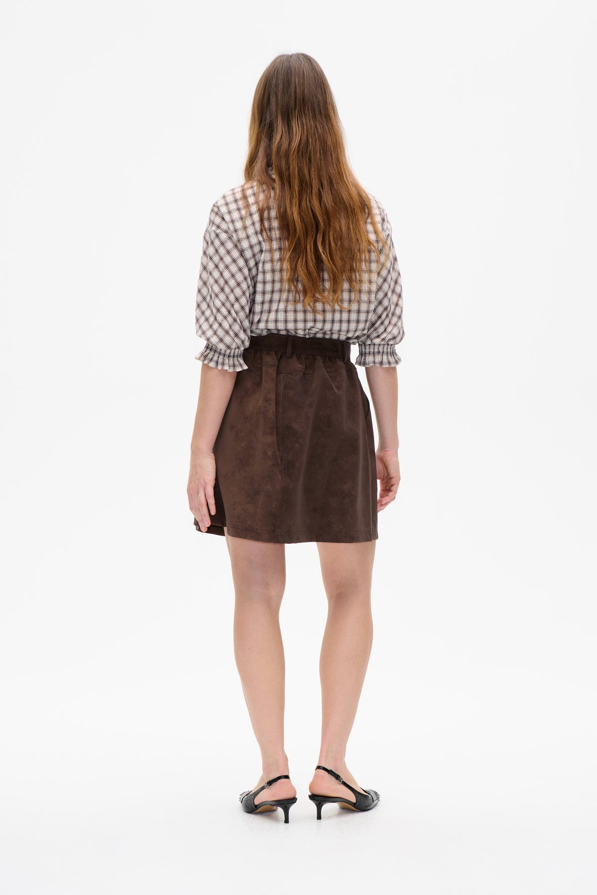 Rear view of a model wearing a cream plaid blouse with voluminous sleeves and ruffled cuffs, paired with a dark brown ruffled mini skirt.