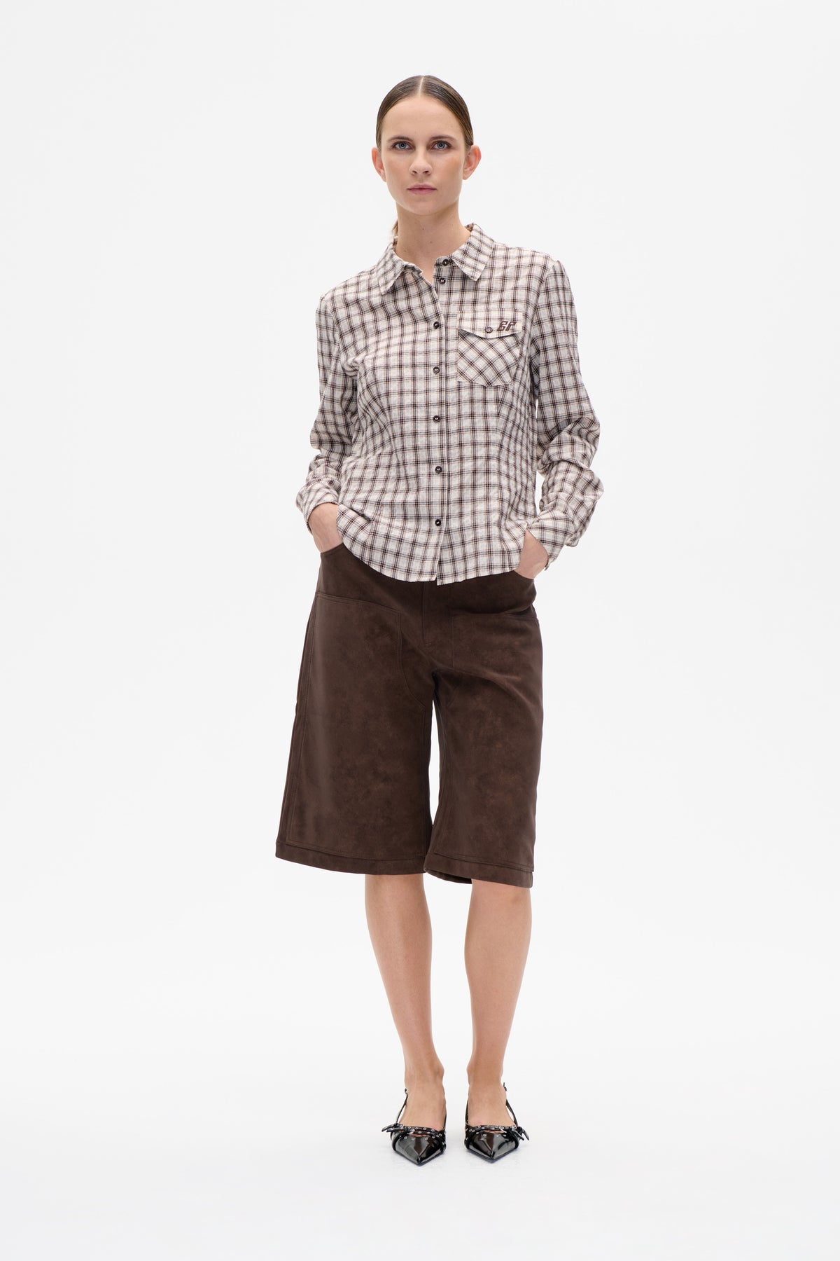 Front view of a model wearing a cream and brown plaid button-front shirt with chest pockets and dark brown wide-leg shorts.