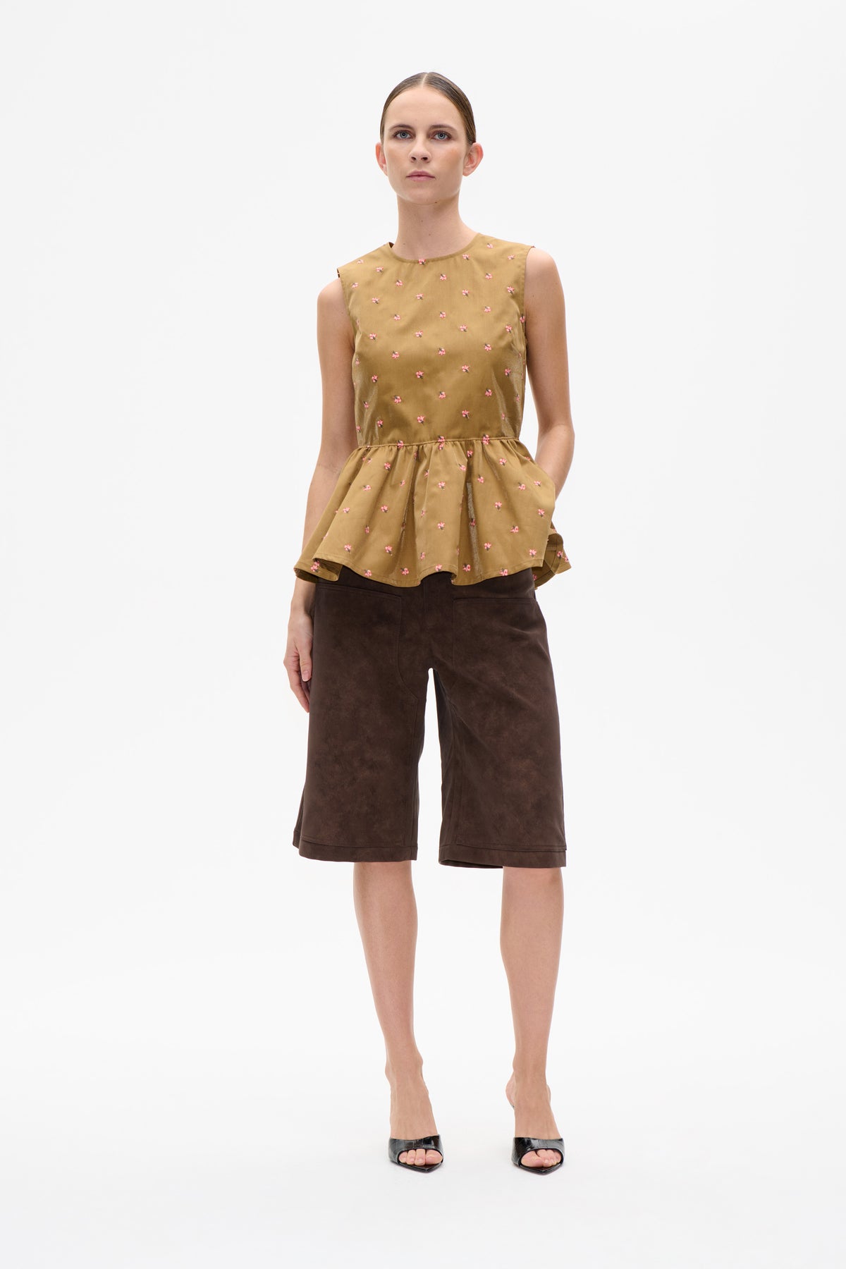 Vorderansicht eines Models, das ein hellbraunes, ärmelloses Peplum-Top mit kleinen rosa Blumen und dunkelbraune, knielange Shorts trägt.