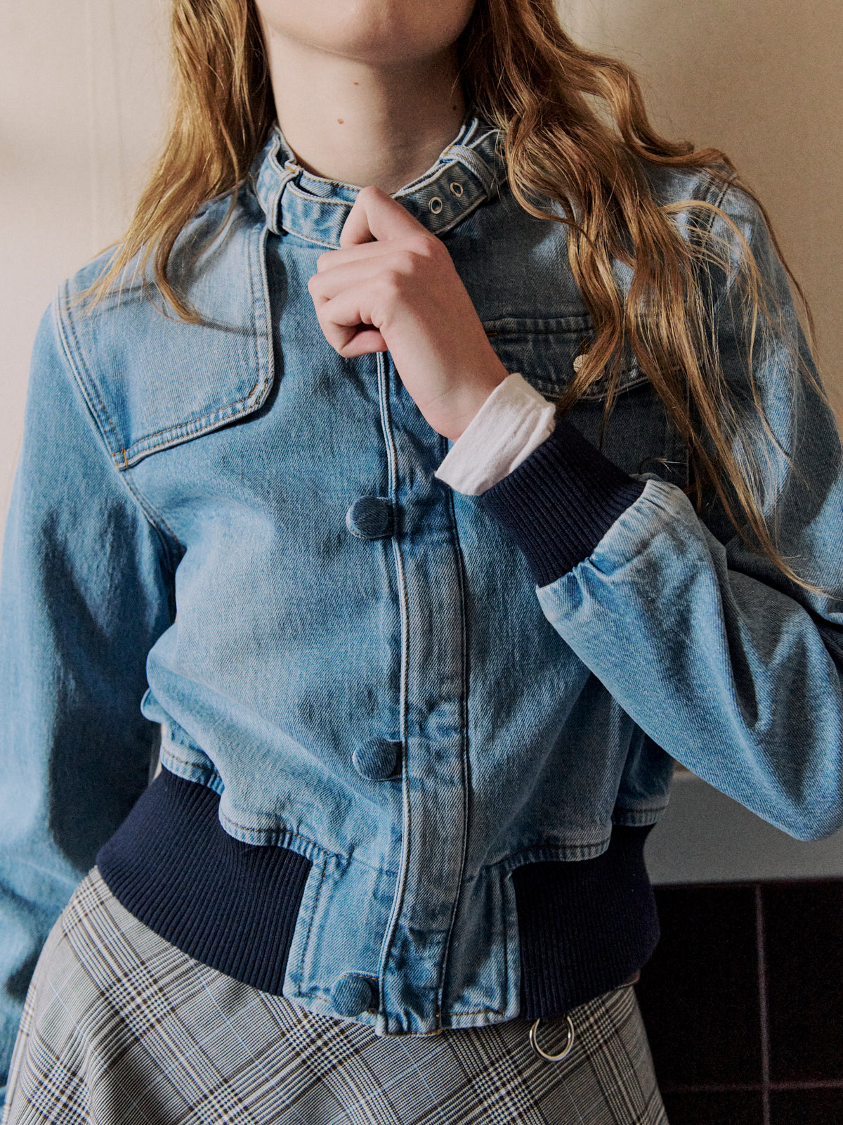 Close-up of a model holding the collar of a light blue denim jacket with oversized covered buttons and navy ribbed cuffs, over plaid trousers.