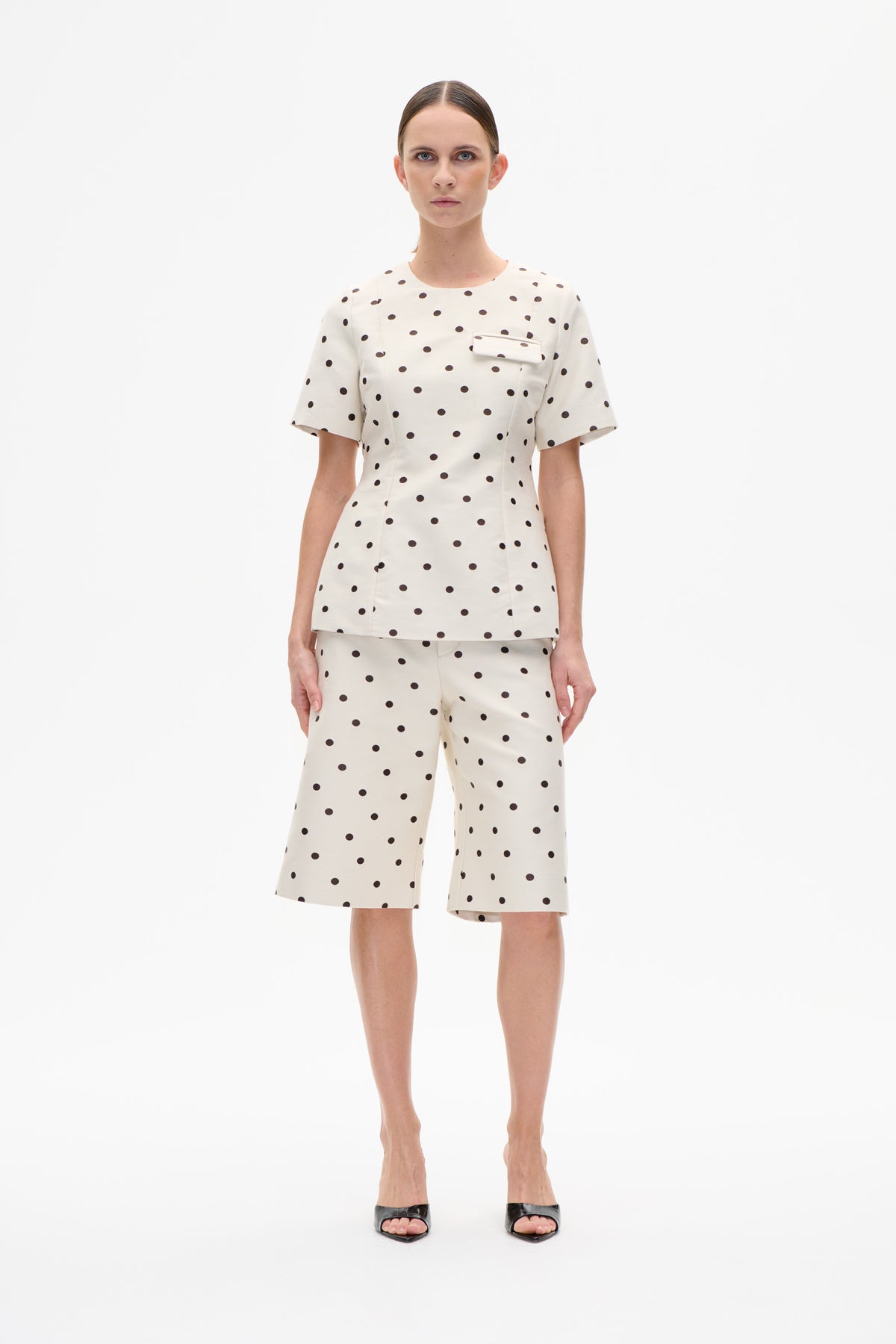 Front view of a model wearing a cream short-sleeved top with a chest flap and matching knee-length shorts, both patterned with dark polka dots.