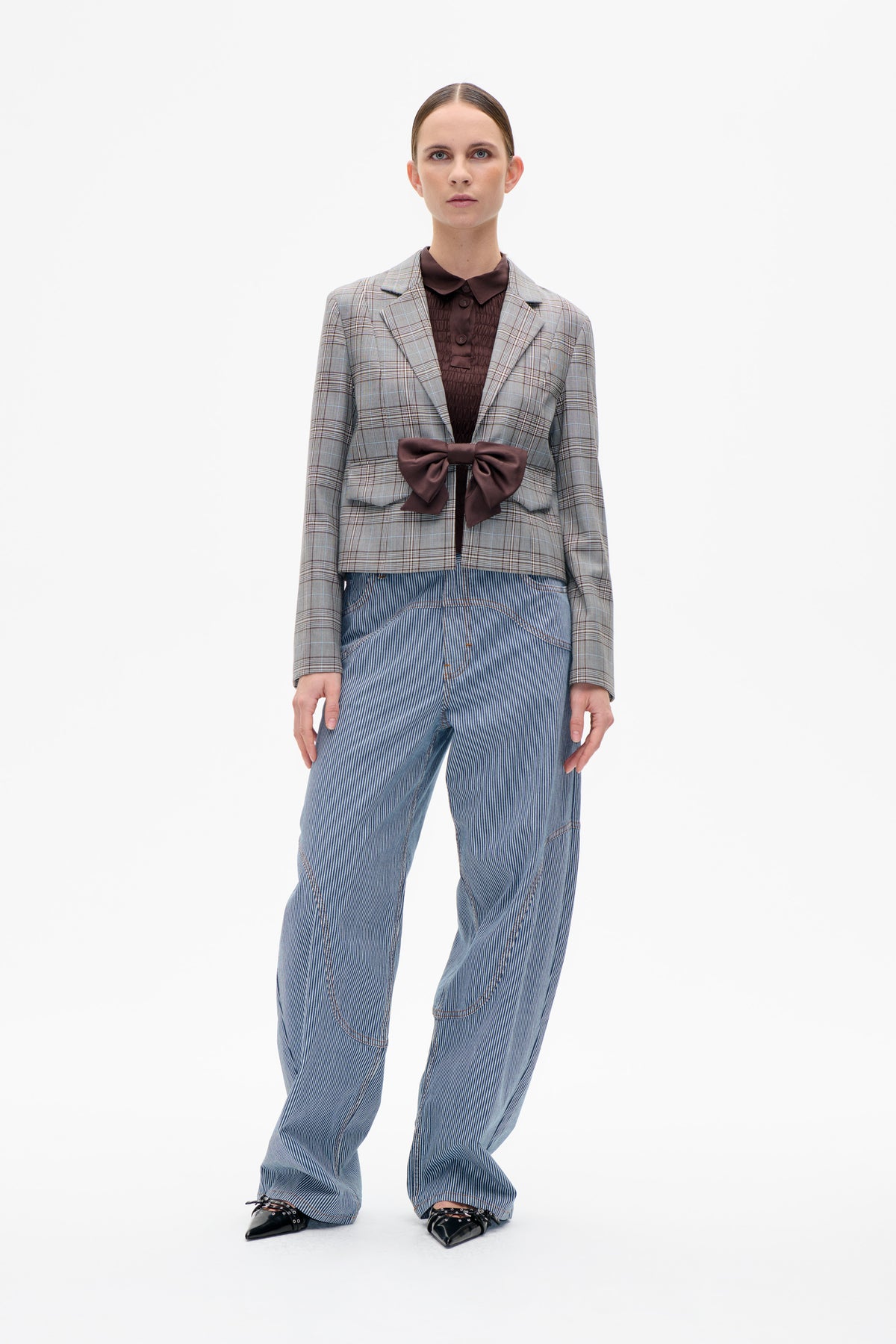 Front view of a model wearing a cropped grey plaid blazer with a large plum bow, a plum quilted shirt and blue pinstriped jeans.