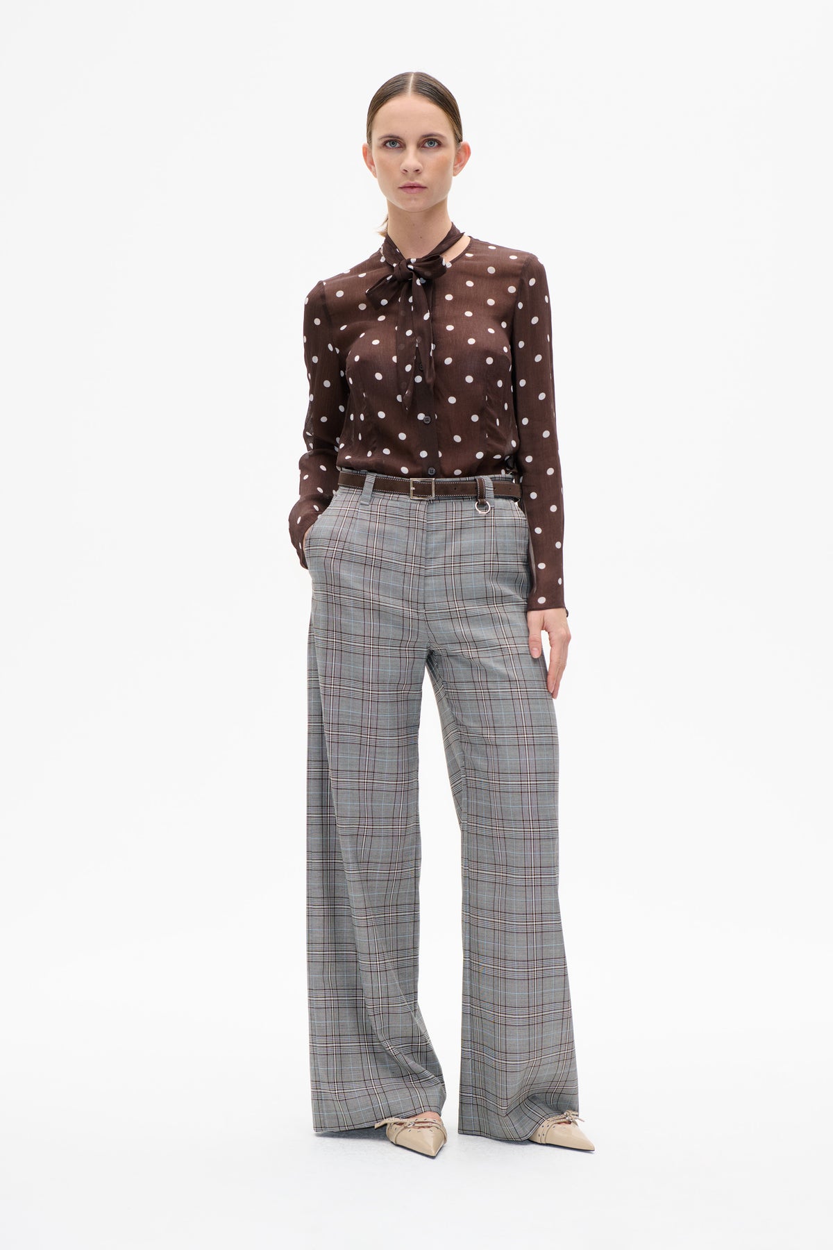 Front view of a model wearing a brown blouse with white polka dots and a neck tie, paired with wide-leg grey plaid trousers.