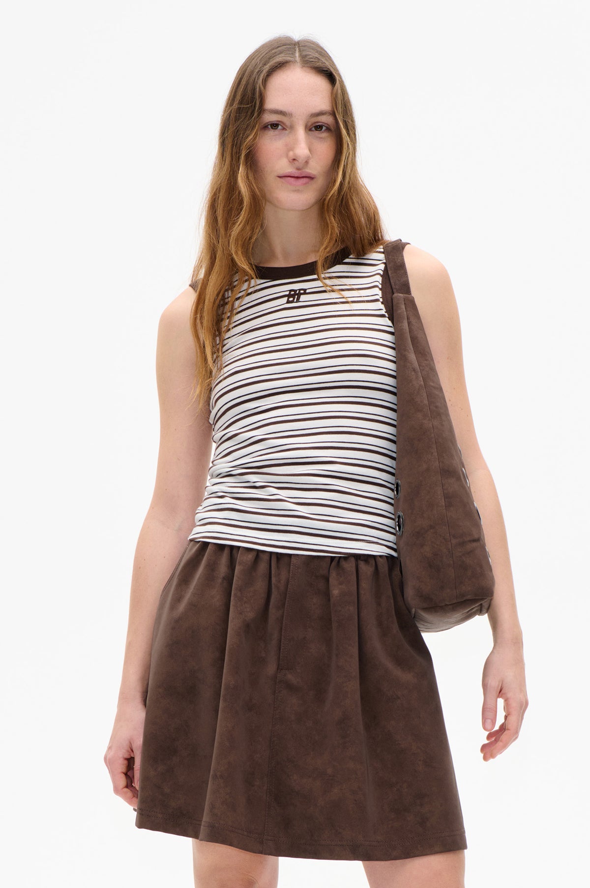 Front view of a model wearing a sleeveless white top with thin dark stripes and a small chest logo, paired with a brown skirt and shoulder bag.