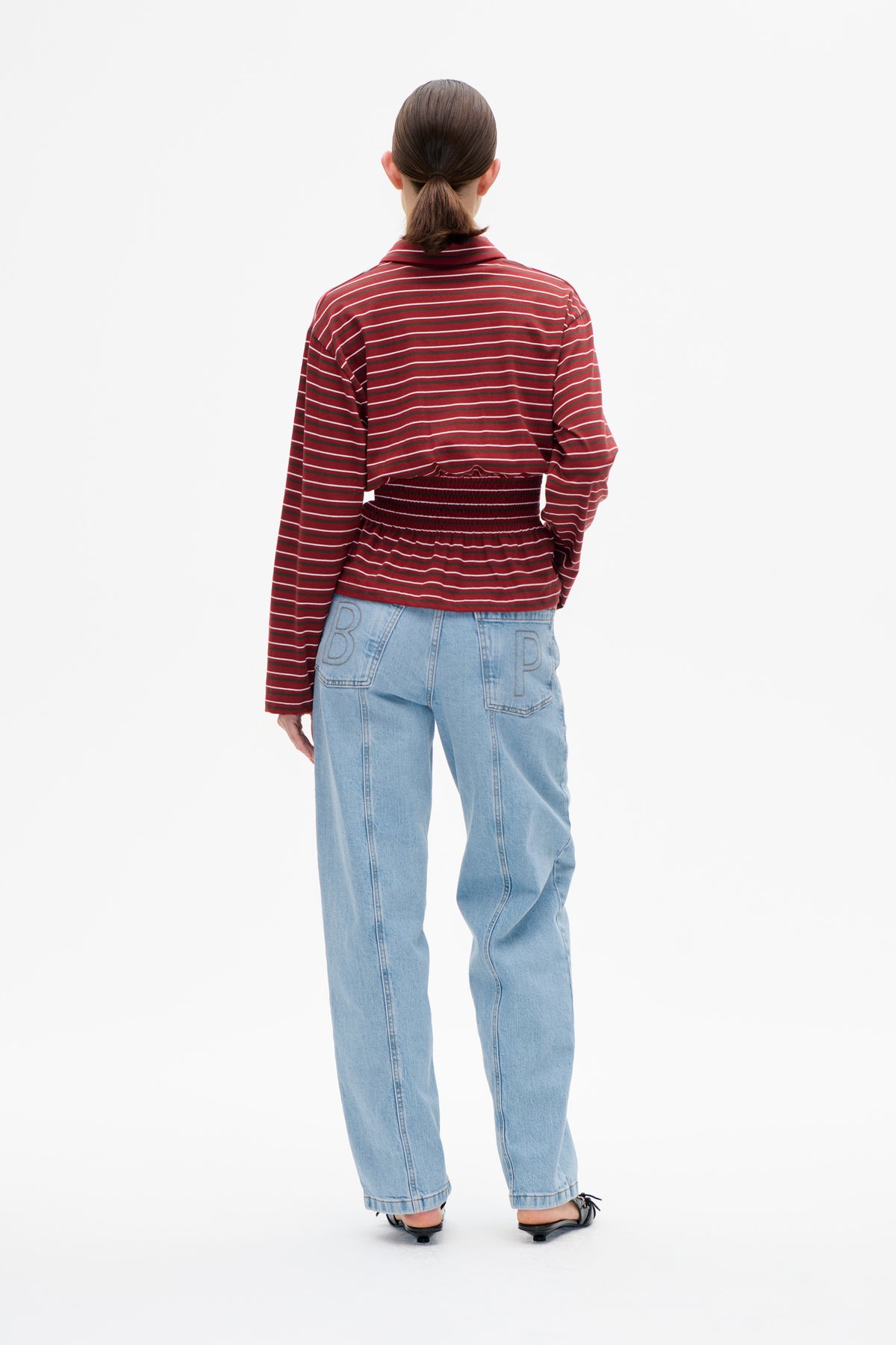 Back view of a model wearing a burgundy-red long-sleeve collared top with thin white and dark stripes and a smocked waist, paired with light-wash jeans.
