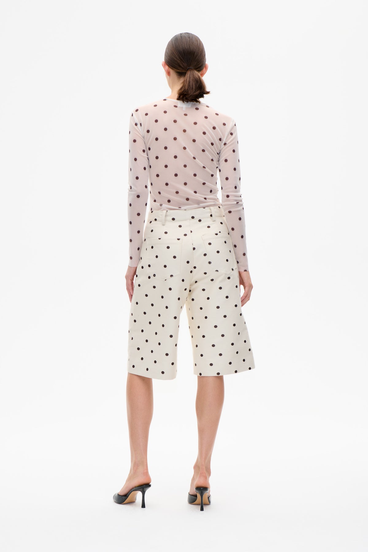 Back view of a model wearing a pale pink long-sleeved top and cream wide-leg shorts, both patterned with dark brown polka dots.