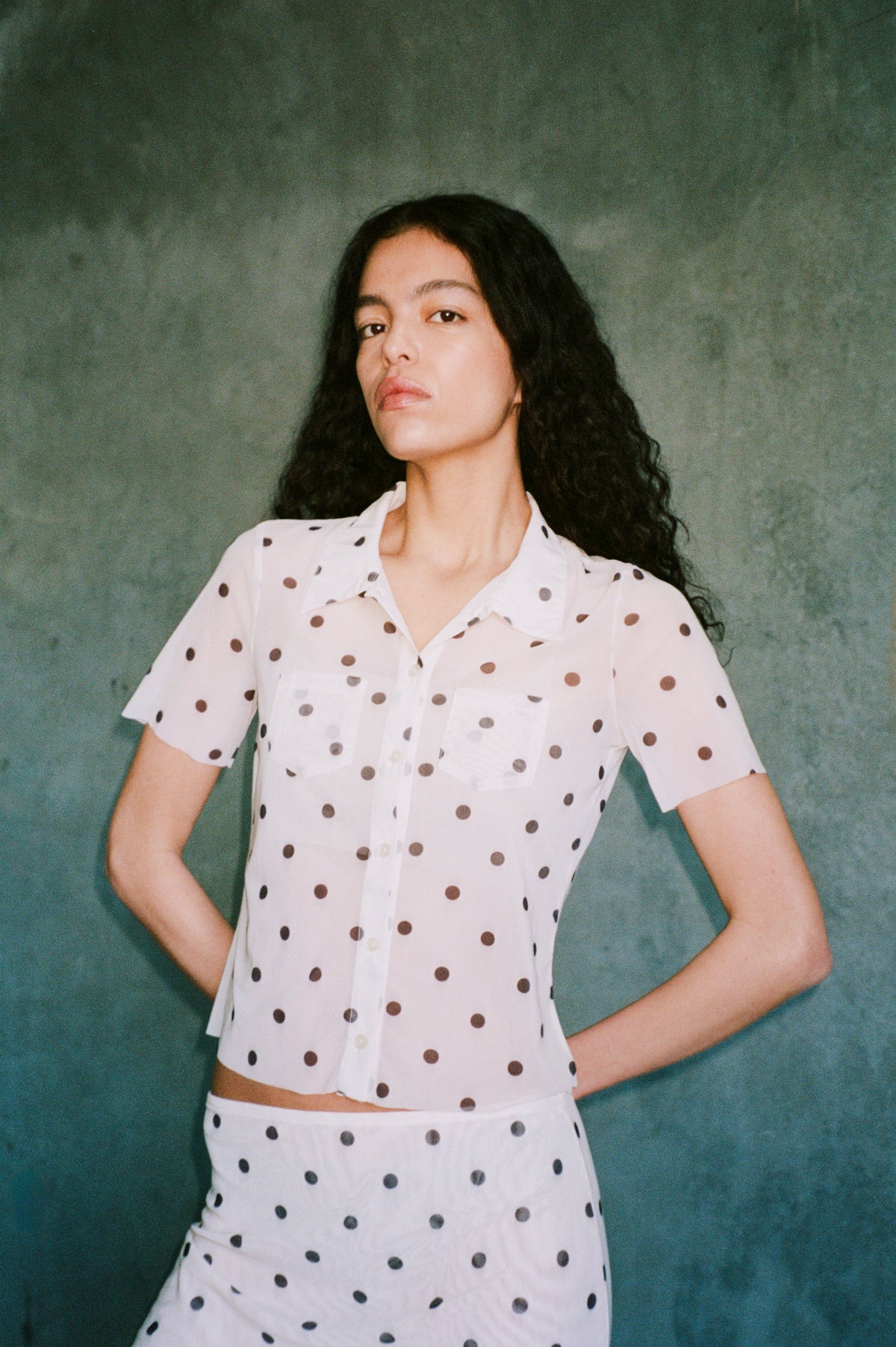 Campaign portrait of a model wearing a white short-sleeved button-front shirt and matching skirt with scattered dark polka dots.
