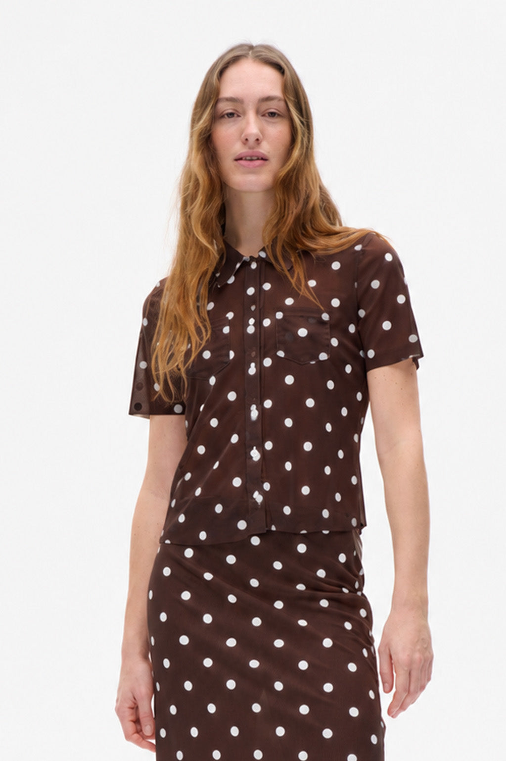 Model draagt een donkerbruine blouse met korte mouwen en knoopsluiting, en een bijpassende rok met witte stippen.
