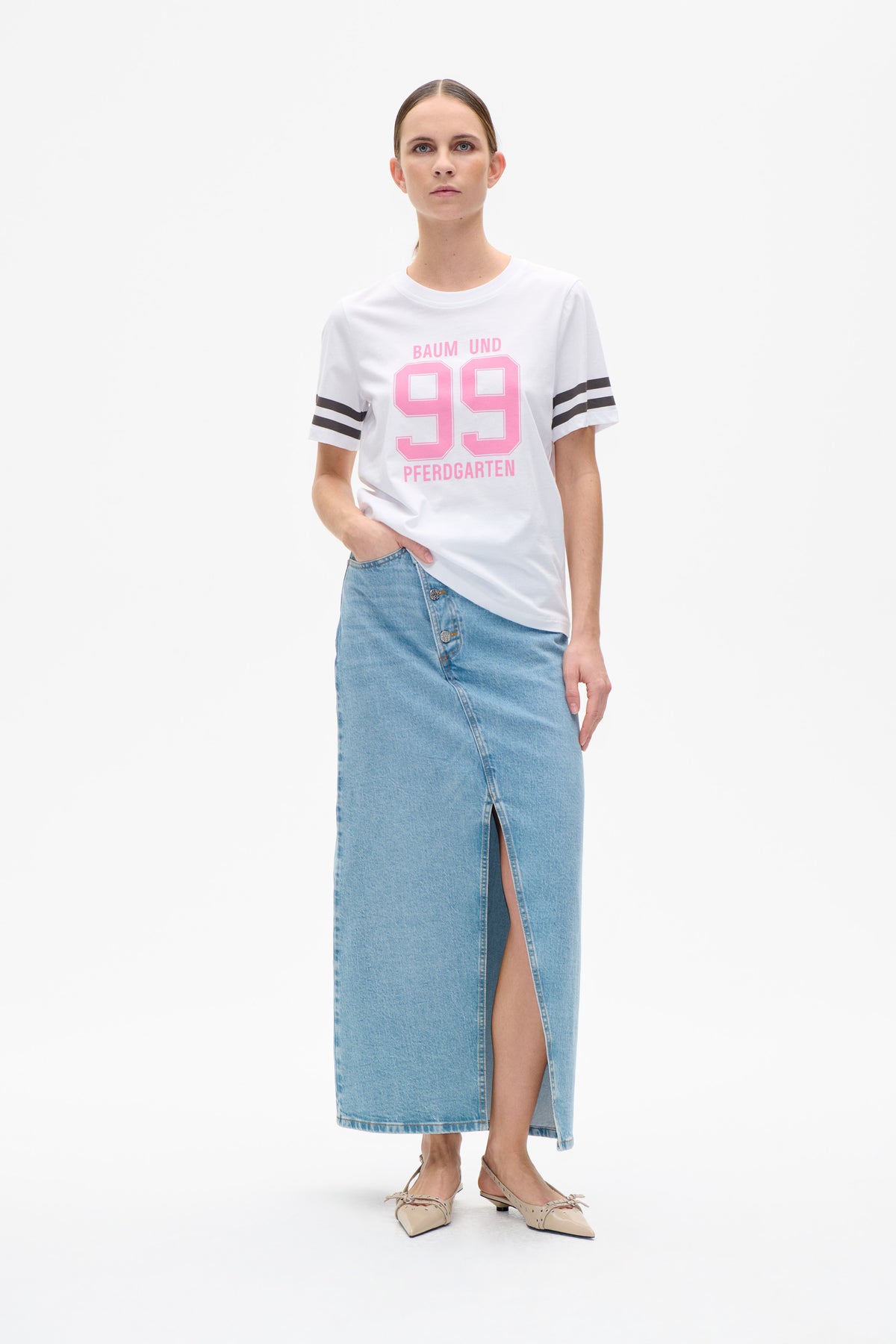 Model wearing a white T-shirt with pink “99” and Baum und Pferdgarten lettering, black sleeve stripes, and a light-blue denim midi skirt with a front slit.