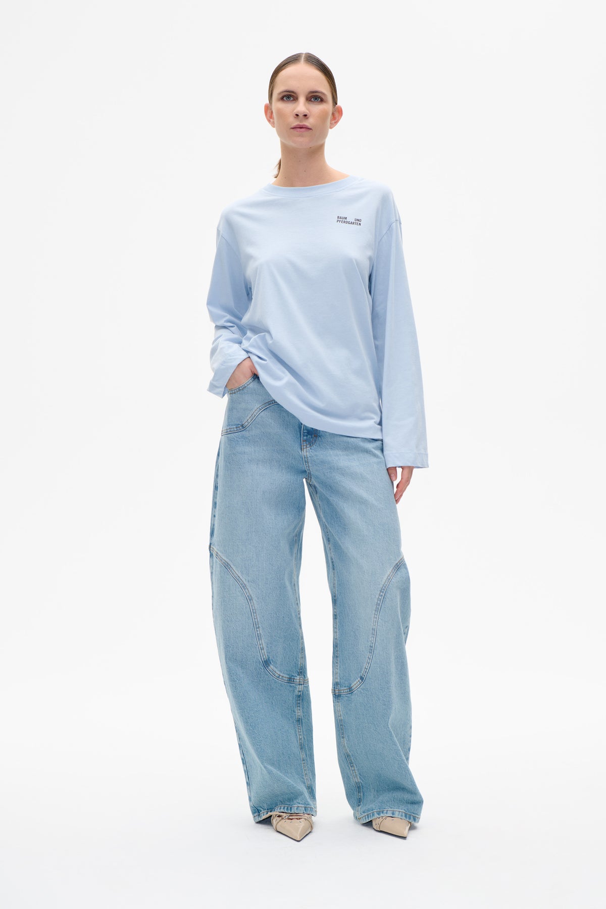 Vorderansicht eines Models, das ein blassblaues Langarm‑T‑Shirt im oversized‑Schnitt mit kleinem Baum und Pferdgarten‑Logo auf der Brust und weit geschnittene, hell gewaschene Jeans trägt.