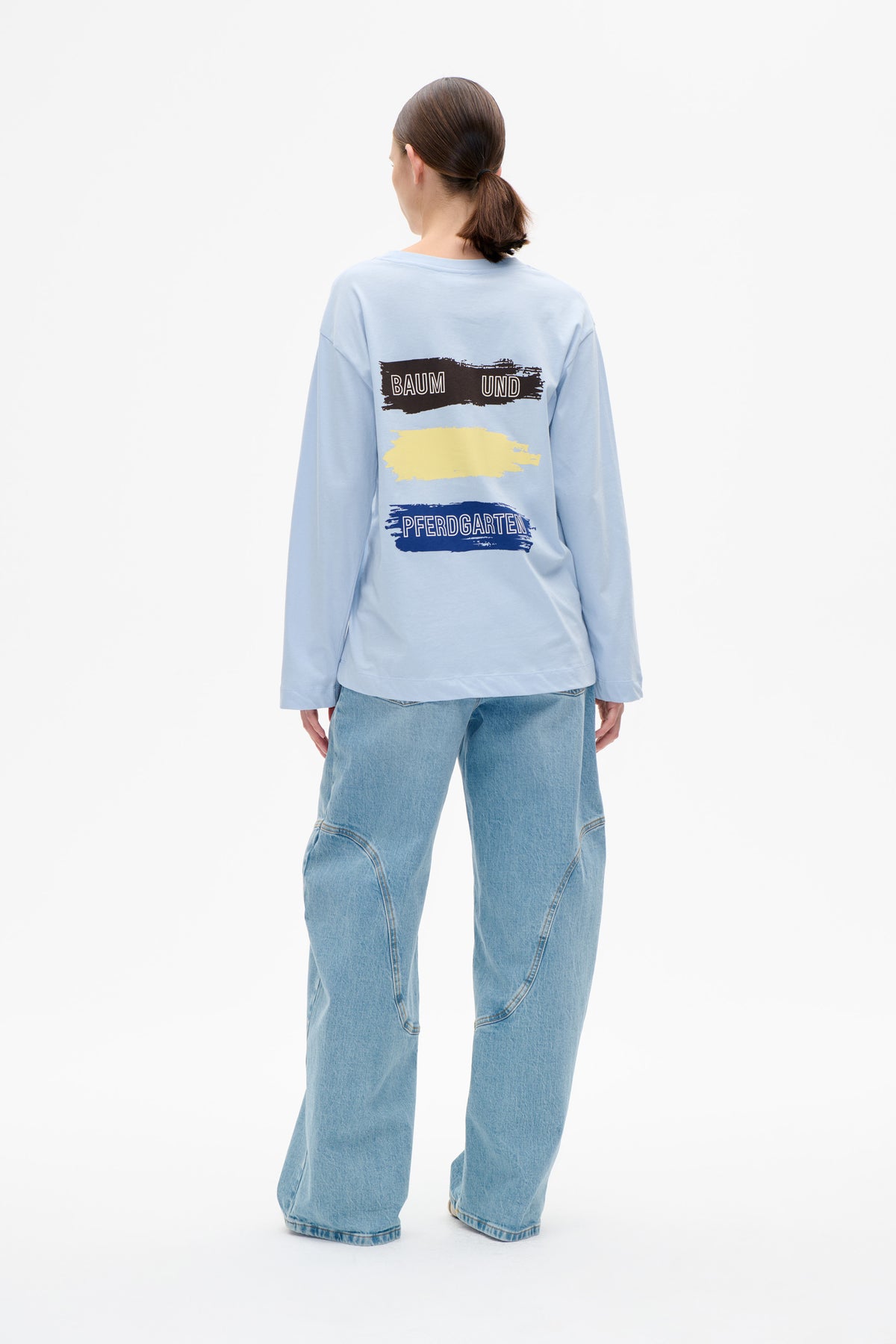 Rückansicht eines hellblauen Langarm-T-Shirts mit Pinselstrich-Motiv, das Baum und Pferdgarten in Schwarz und Marineblau zeigt, mit einem gelben Streifen.