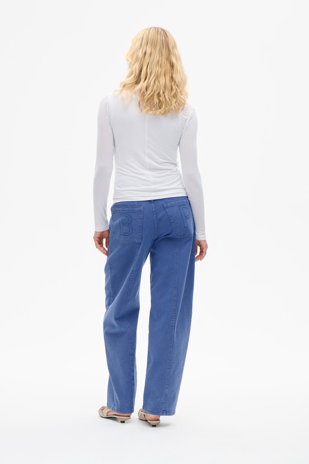 Back view of a model wearing a fitted white long-sleeve top and blue jeans with outlined lettering on the rear pockets.