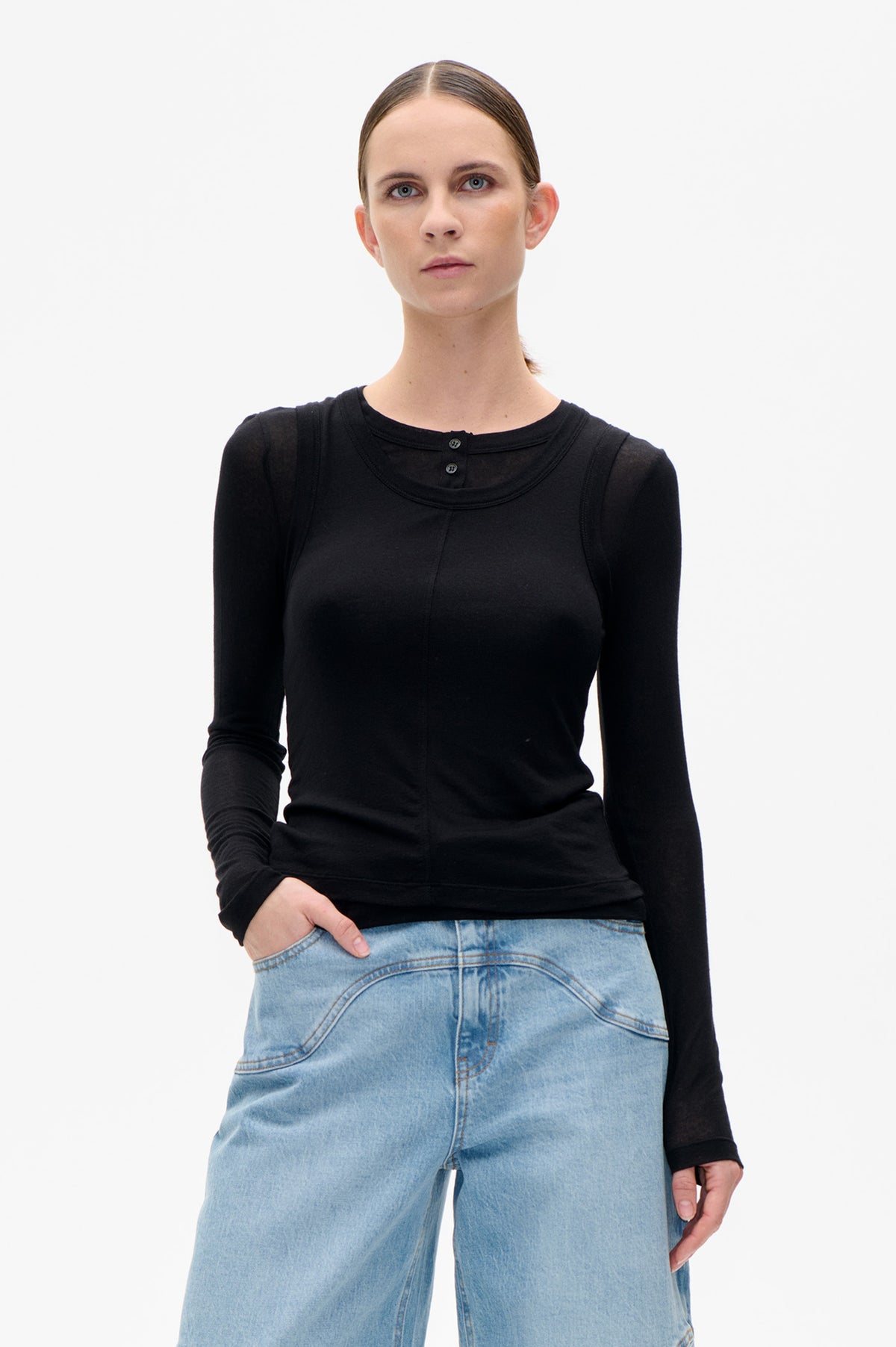 Model wearing a black long-sleeve top with a rounded neckline, two buttons and visible seam detailing, styled with light-wash jeans.