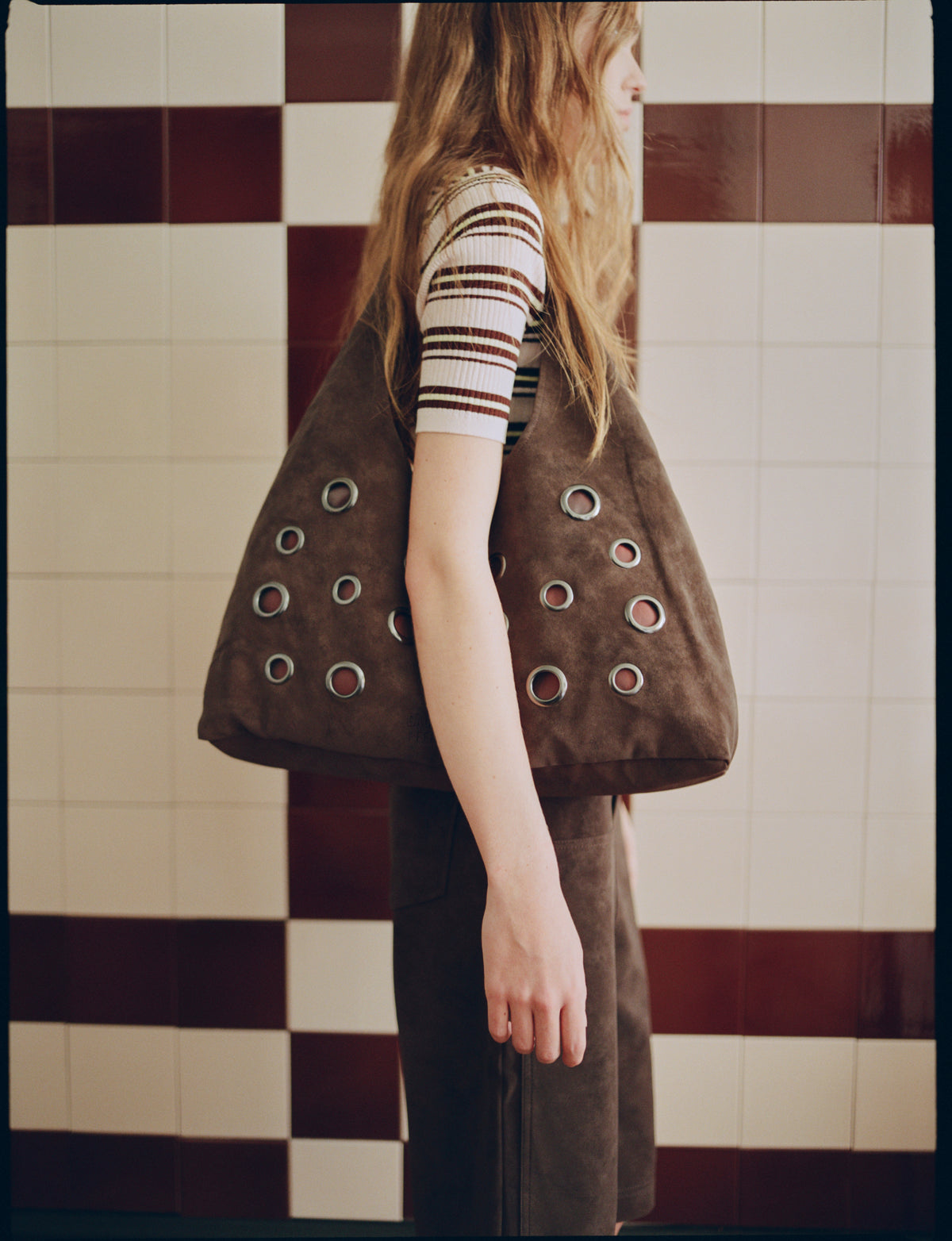 Side view of a model carrying an oversized brown bag with metal eyelets, wearing a white ribbed top with burgundy stripes and brown trousers.
