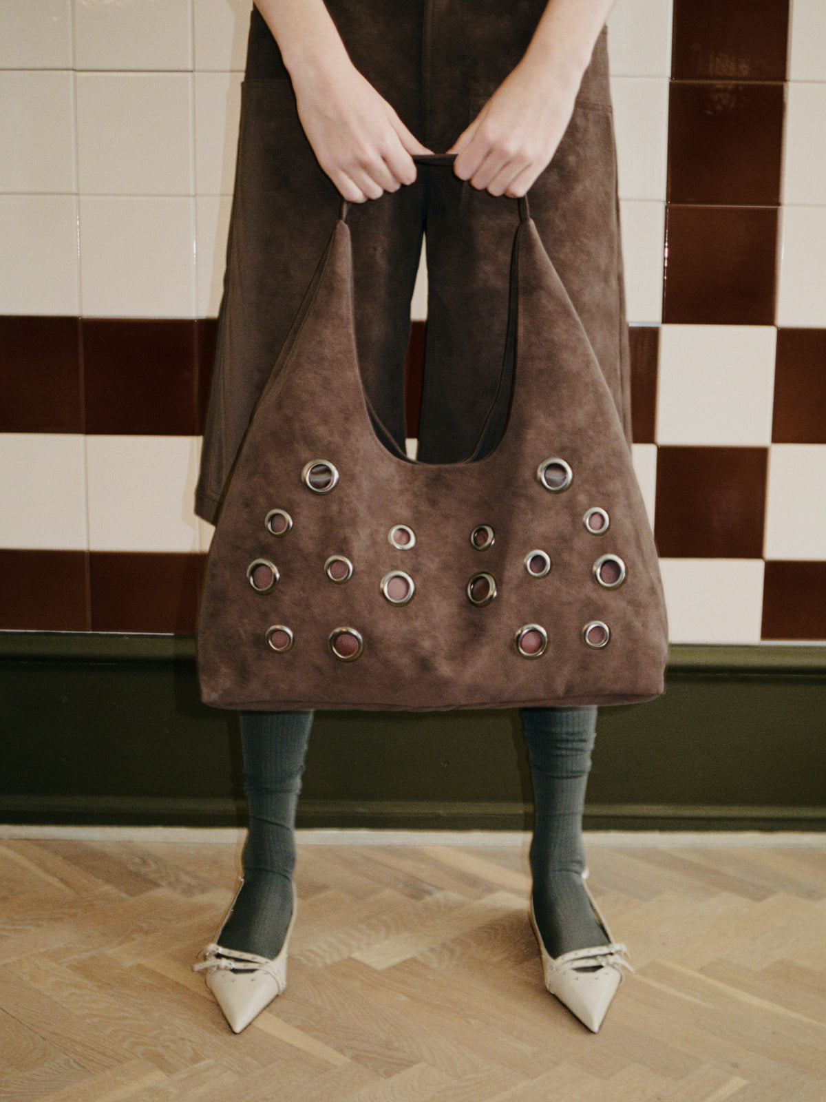Model holds a large taupe-brown tote with silver eyelet details, styled with grey tights and white pointed shoes against tiled walls.