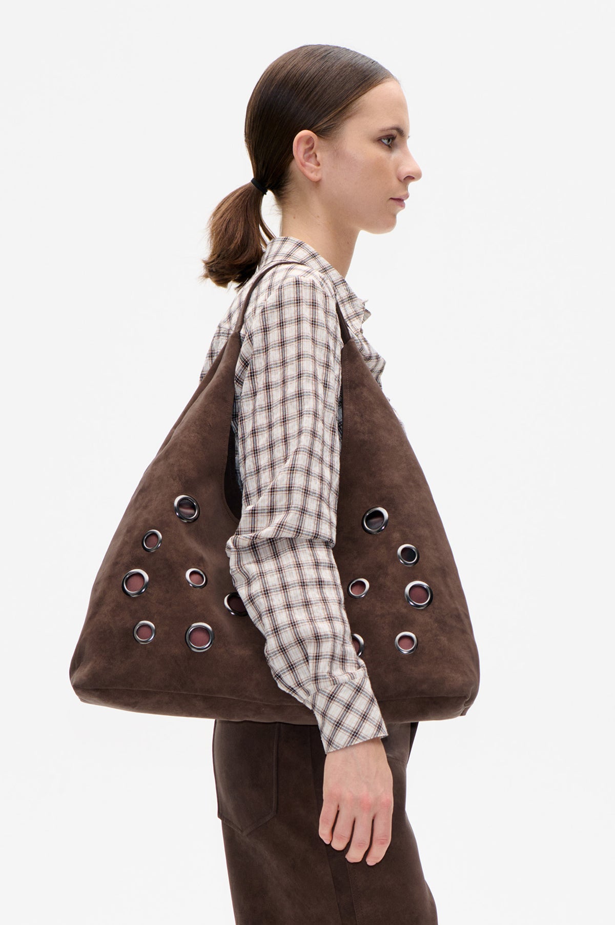 Model in side profile carrying an oversized brown tote with metal eyelets, wearing a cream and brown plaid shirt.