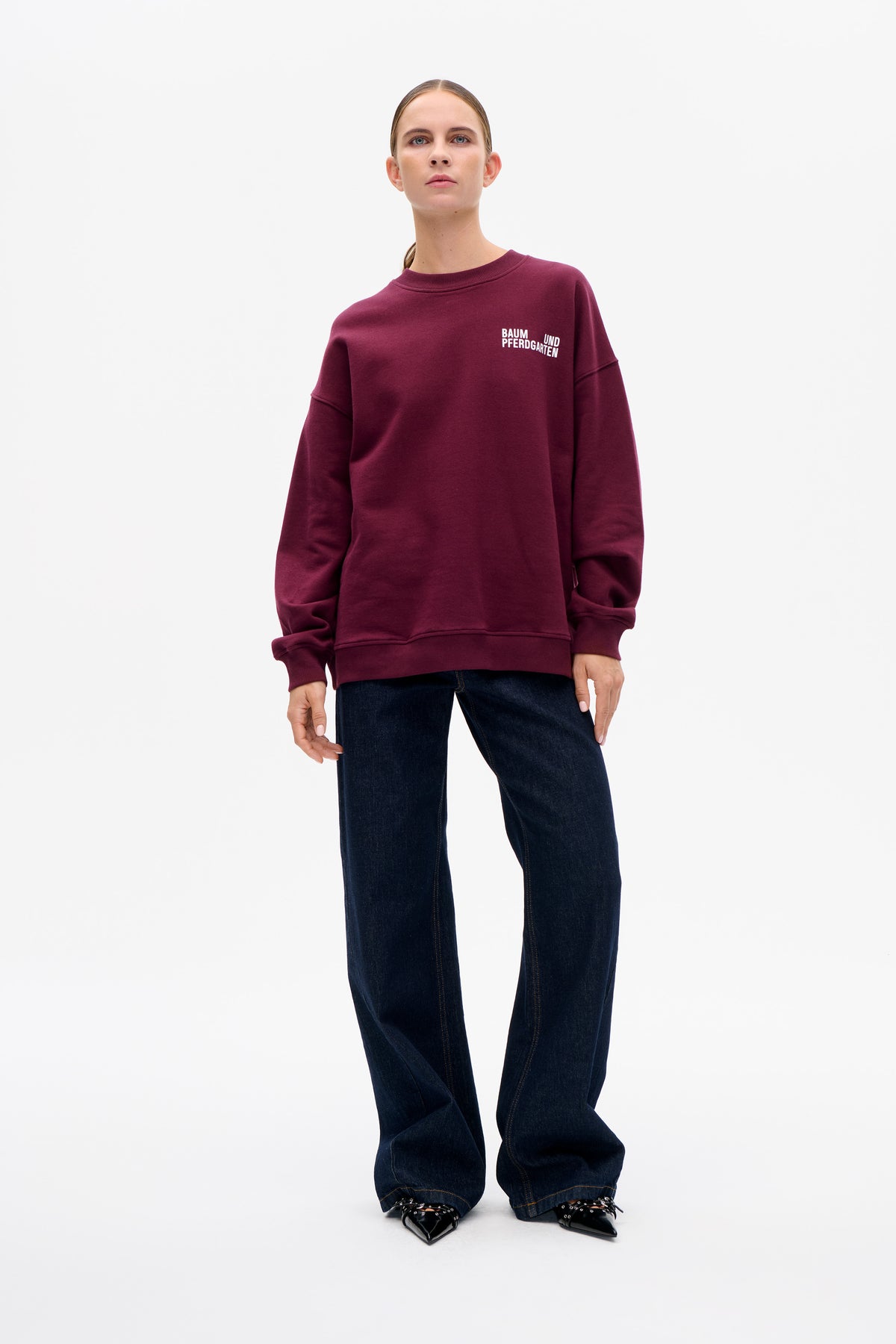 Model draagt een bordeauxrode trui met ronde hals en een klein wit Baum und Pferdgarten borstlogo, donkere wide-leg jeans en zwarte puntige schoenen.